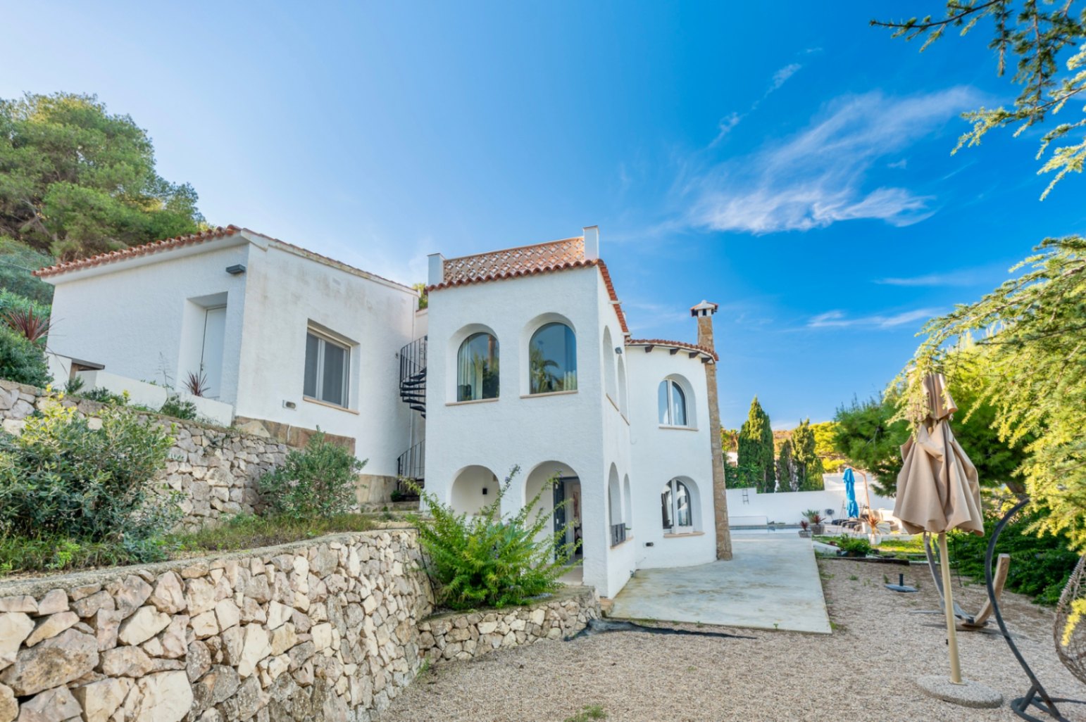 Mediterrane Außenansicht der Villa mit Rundbogenfenstern, einer Wendeltreppe zur Dachterrasse und Trockensteinmauern in Hanglage.