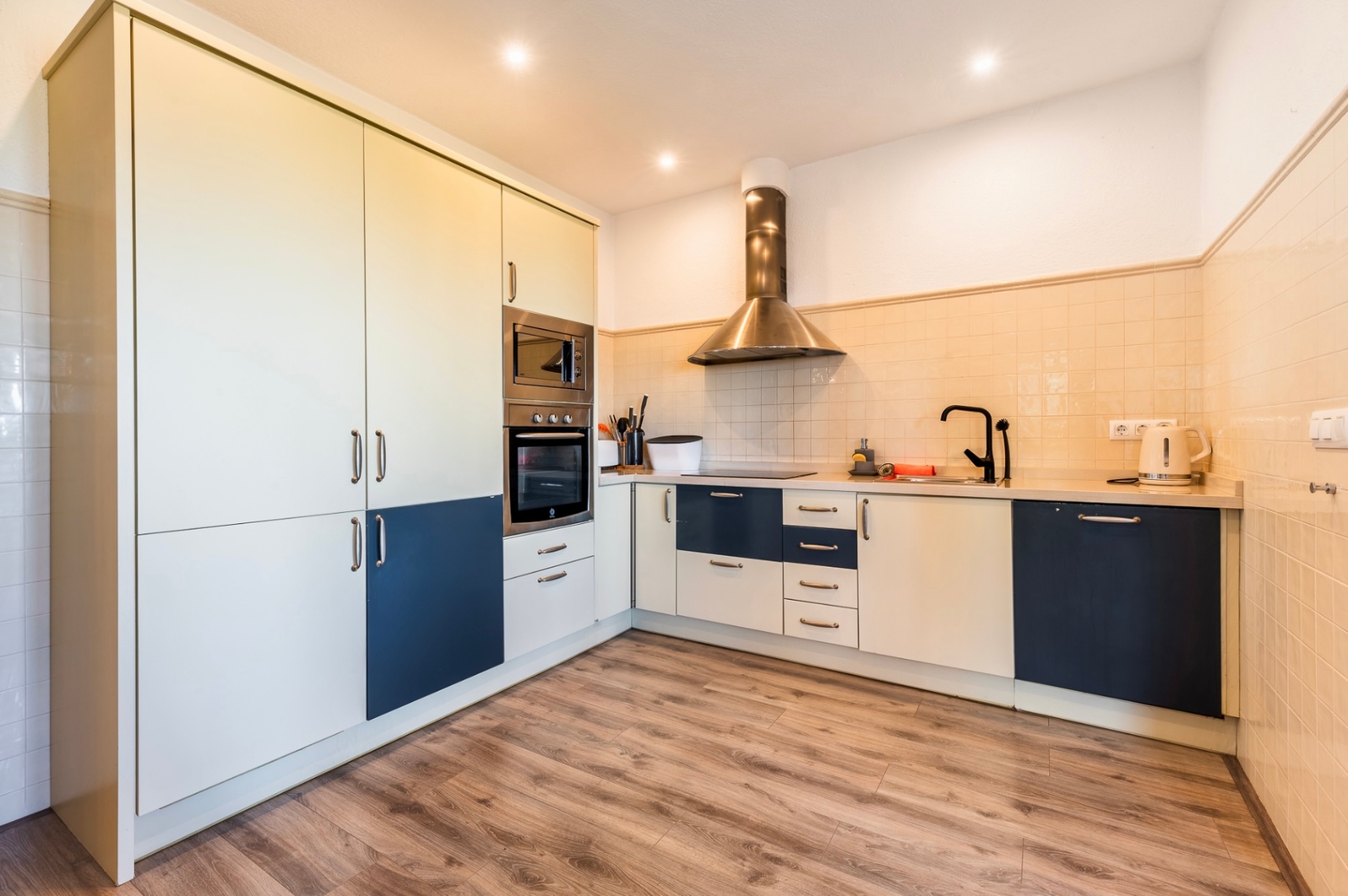 Moderne Küche mit zweifarbigen Schränken in Creme und Blau, integrierten Edelstahlgeräten, einer Dunstabzugshaube und Laminatboden in Holzoptik.