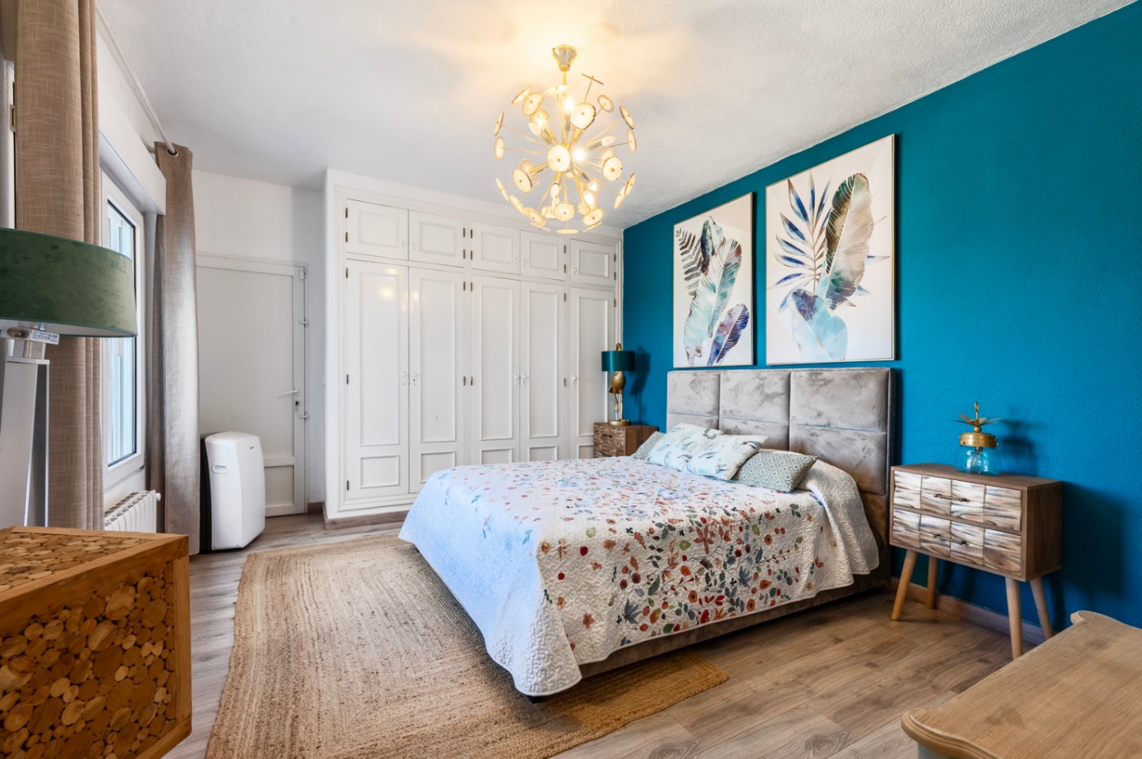 Schlafzimmer mit türkisfarbener Akzentwand, weißen Einbauschränken und gepolstertem Kopfteil. Mit Laminatboden, Jute-Teppich und einem modernen goldenen Kronleuchter.