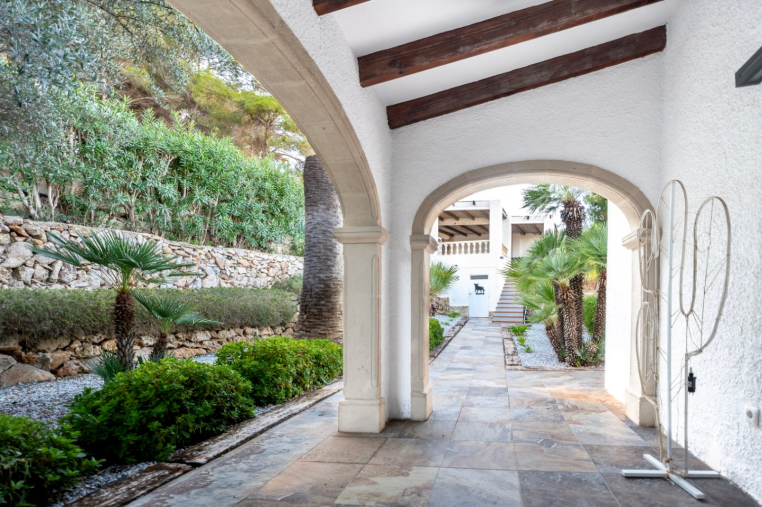 Traditionelle Veranda mit Tosca-Steinbögen, sichtbaren Holzbalken und Natursteinböden mit Blick auf den mediterranen Garten und Trockensteinmauern.