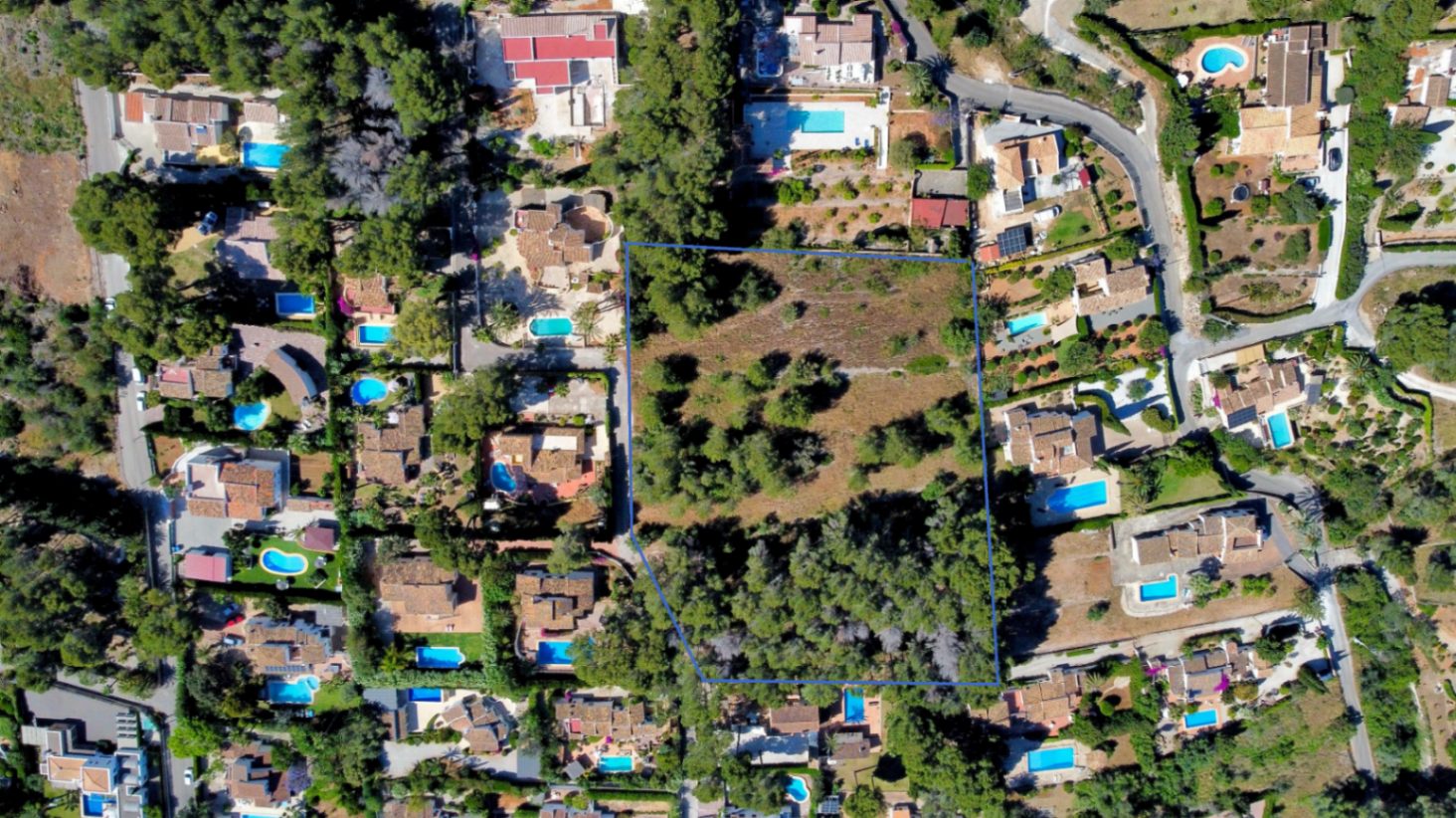 Luftaufnahme des blau umrandeten, flachen Grundstücks, umgeben von Wohnvillen mit Swimmingpools und üppiger mediterraner Vegetation.