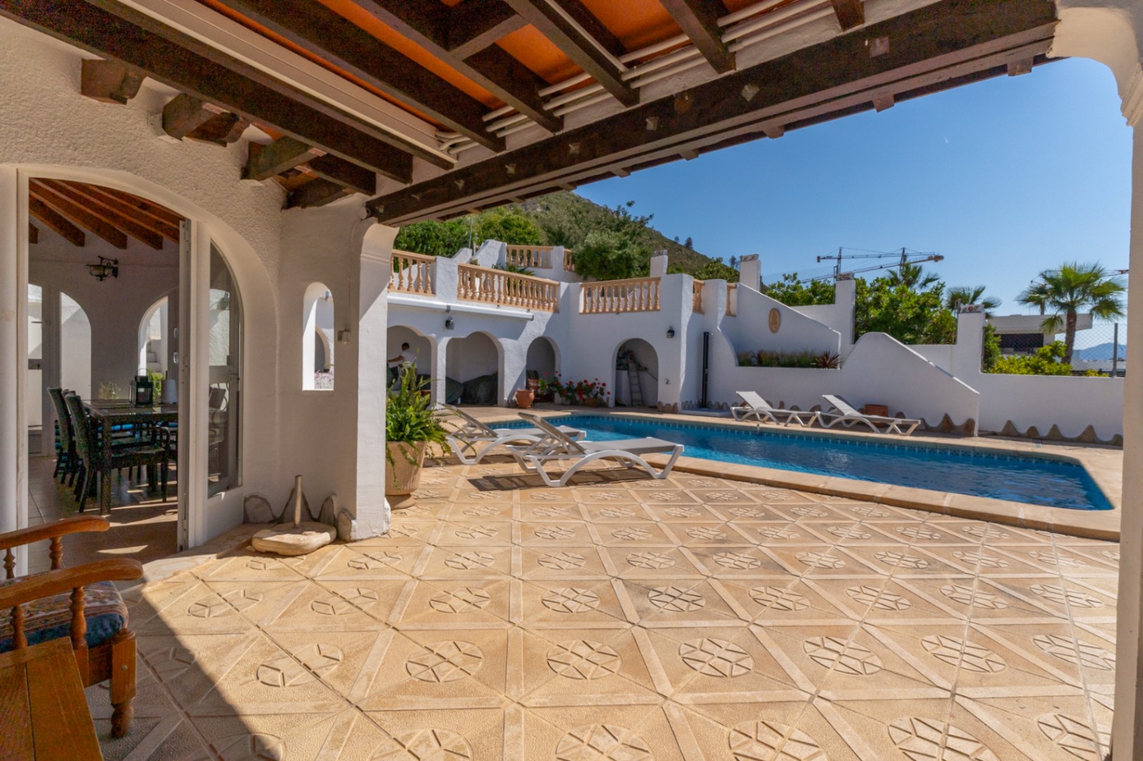 Außenbereich mit gemusterten Steinfliesen, rechteckigem Pool und überdachter Veranda mit sichtbaren Holzbalken und mediterranen Rundbögen.