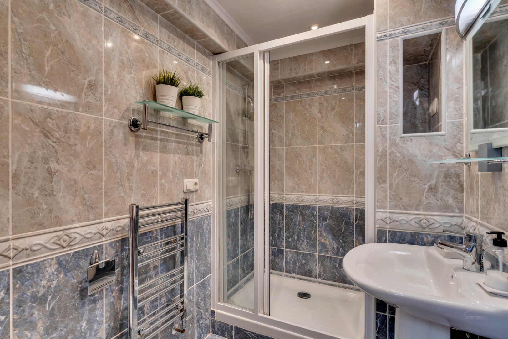 Badezimmer mit Duschkabine, beigen und blauen Fliesen in Marmoroptik, verchromtem Handtuchheizkörper und klassischem Standwaschbecken.