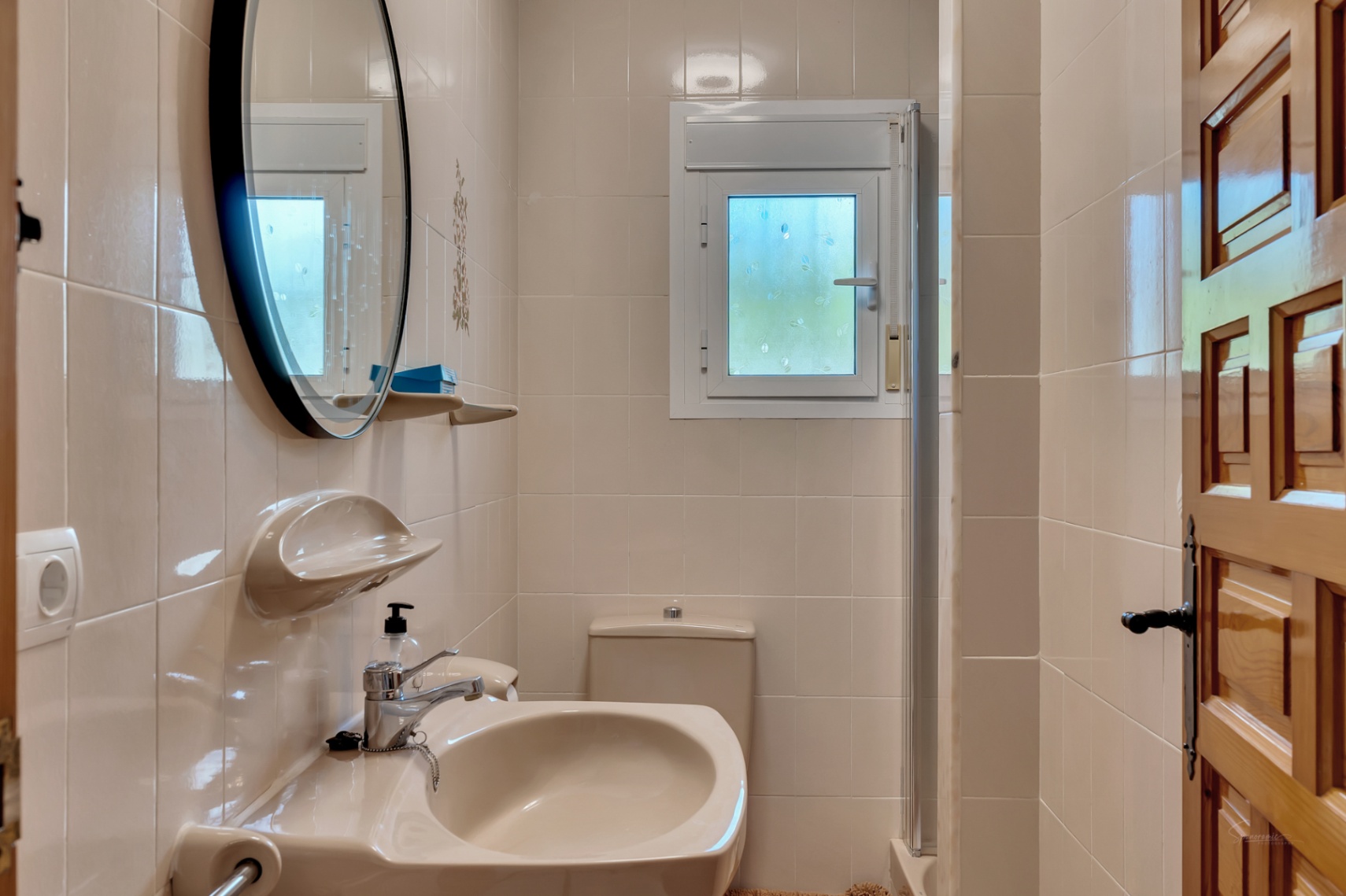 Funktionales Badezimmer mit cremefarbenen Fliesen, rundem Spiegel mit schwarzem Rahmen, Keramikwaschbecken und kleinem Fenster für natürliches Licht.