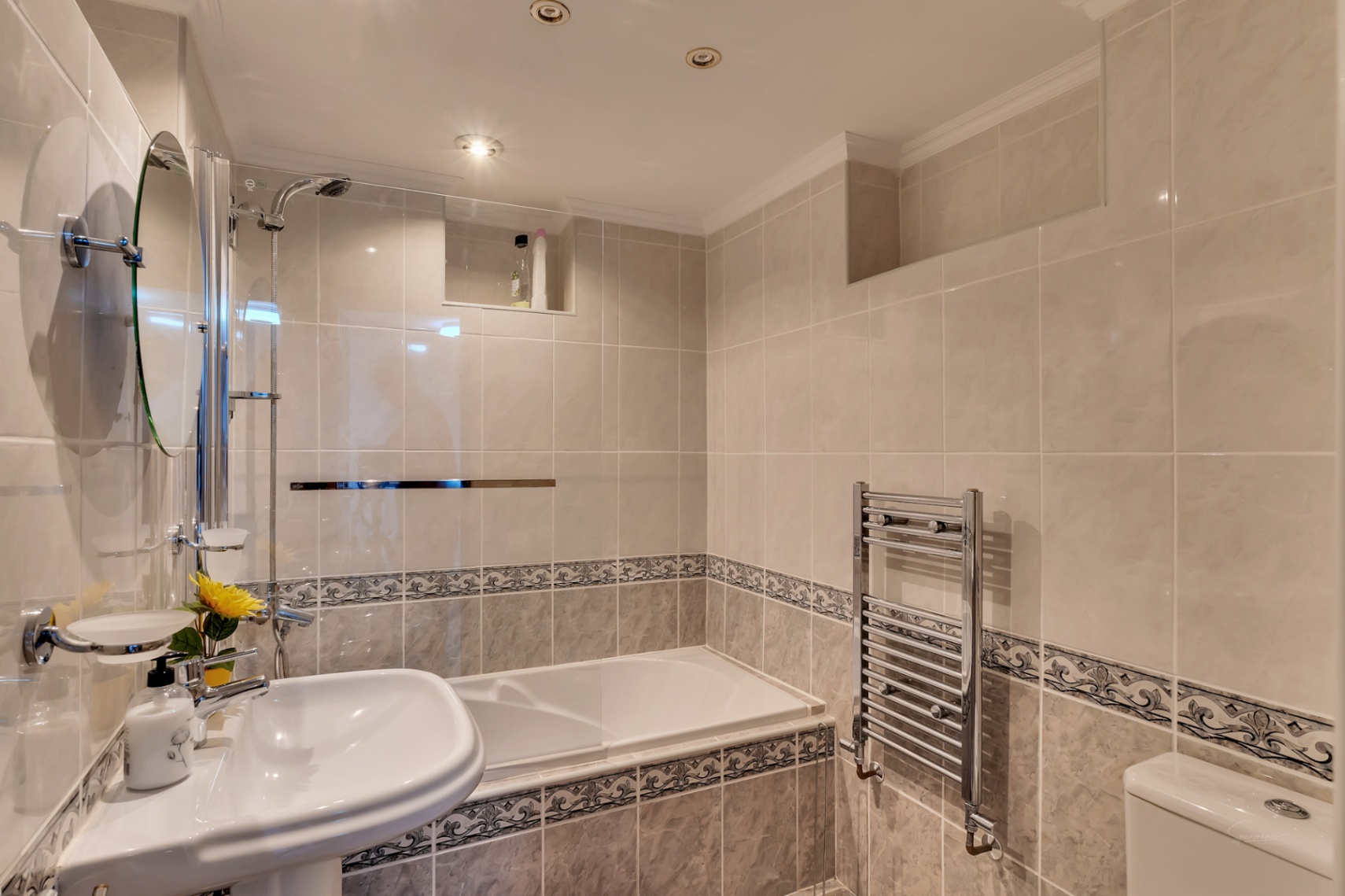 Gefliestes Badezimmer mit Einbaubadewanne, Glasduschwand, Standwaschbecken, verchromtem Handtuchheizkörper und Deckeneinbaustrahlern.