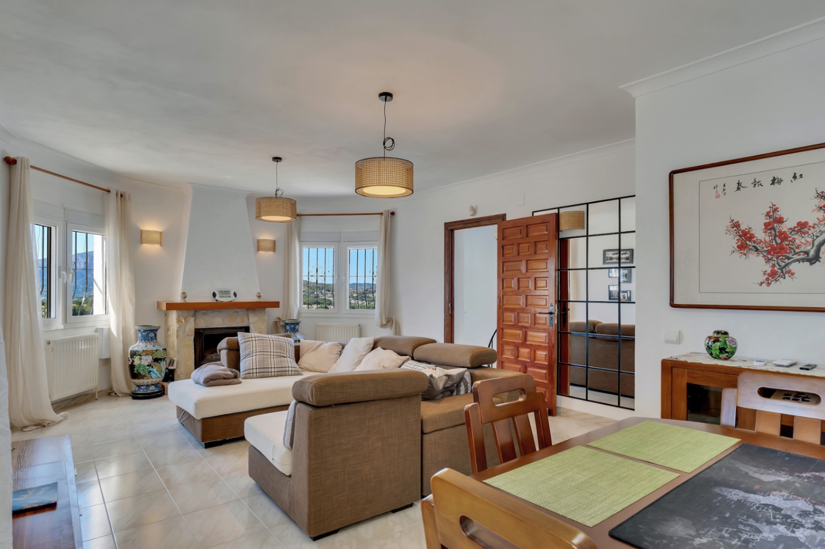 Heller Wohn- und Essbereich mit Natursteinkamin, modularen Sofas und Blick auf den Montgó. Elegante Details wie Rattan-Pendelleuchten, Fliesenboden und ein großer Rasterspeigel.