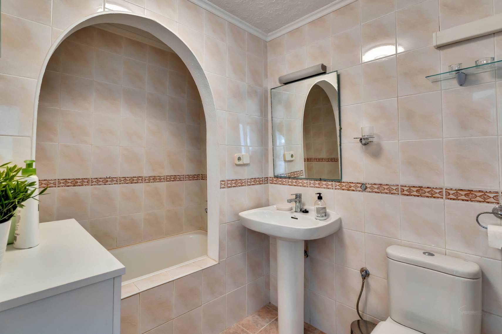 Klassisches Badezimmer mit einer in eine Rundbogennische integrierten Badewanne, Säulenwaschbecken und cremefarbenen Wandfliesen mit Zierborte.