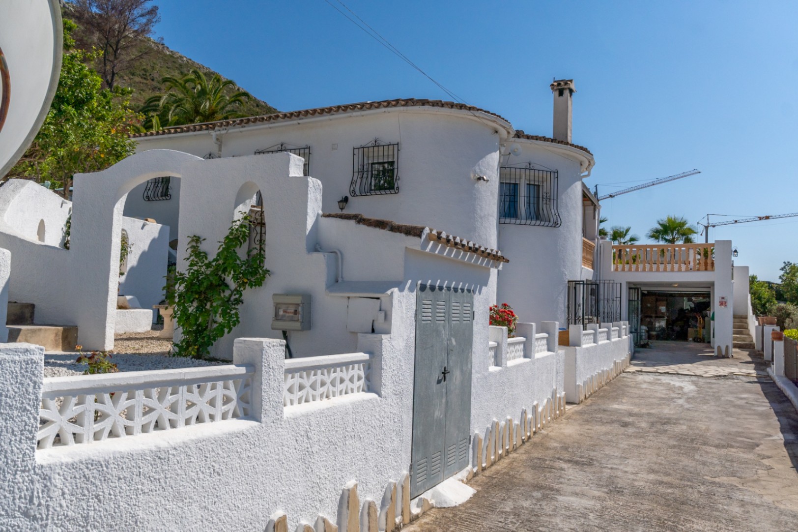 Mediterrane Außenfassade mit weißen Wänden, Rundbögen, verzierten Brüstungen und einer gepflasterten Auffahrt zum Garagenbereich.