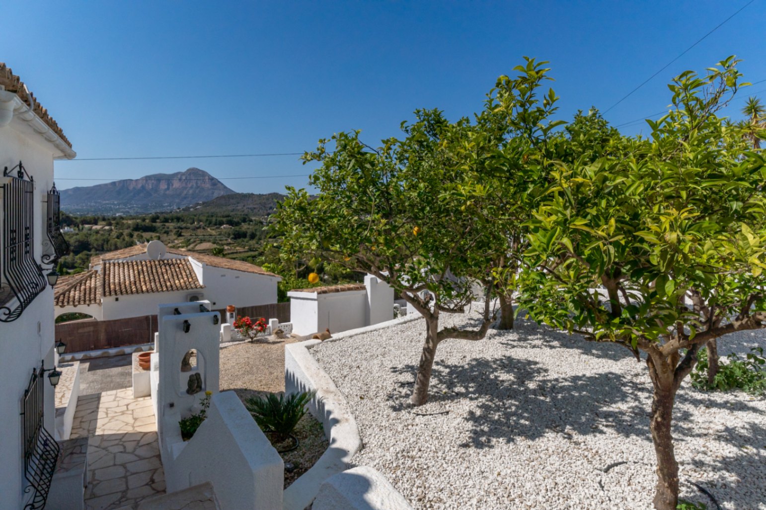 Mediterraner Garten mit weißem Kies, Zitrusbäumen und freiem Blick auf den Montgó. Natursteinweg entlang der traditionellen weißen Architektur der Villa.