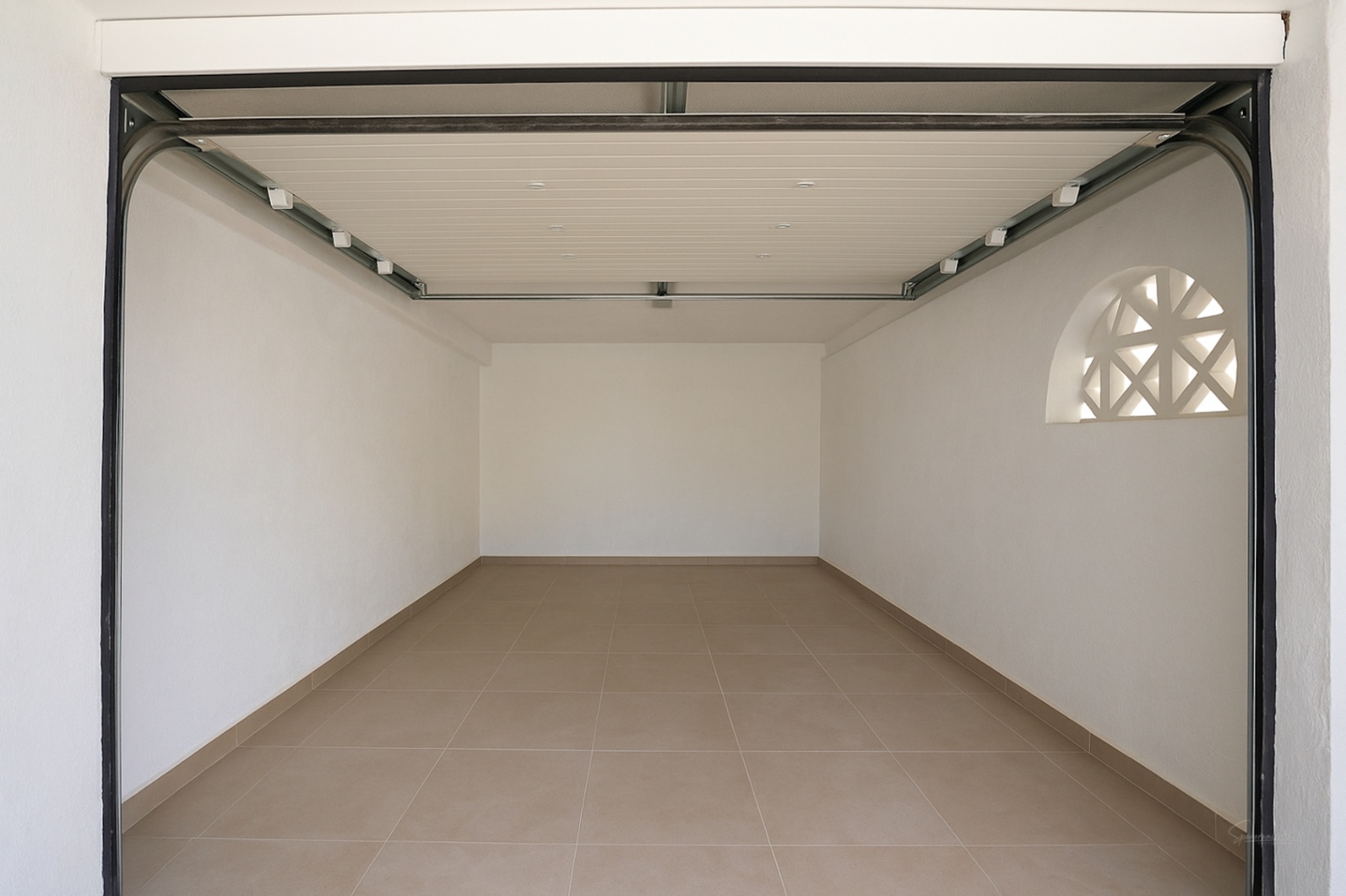 Private Garage mit Fliesenboden, weißen Wänden und automatischem Sektionaltor. Ein dekoratives Rundbogenfenster sorgt für natürliches Licht.