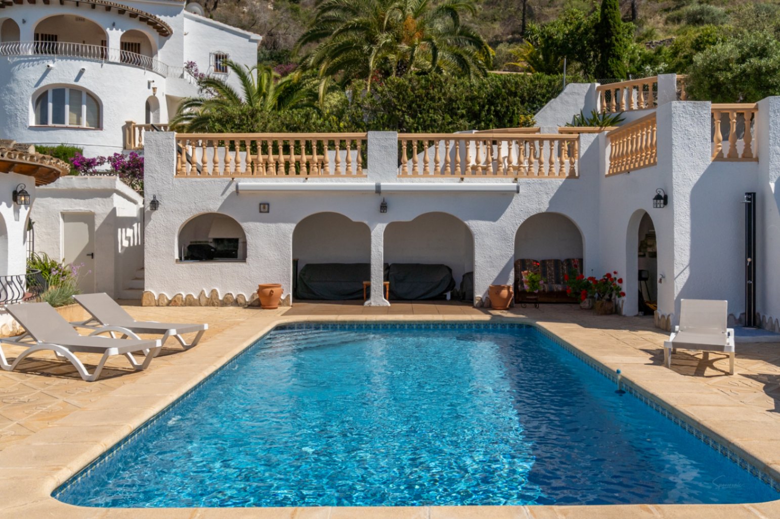 Rechteckiger Pool auf einer Steinterrasse mit Sonnenliegen, Rundbogen-Veranden und klassischen weißen Balustraden vor einer grünen Hügellandschaft.