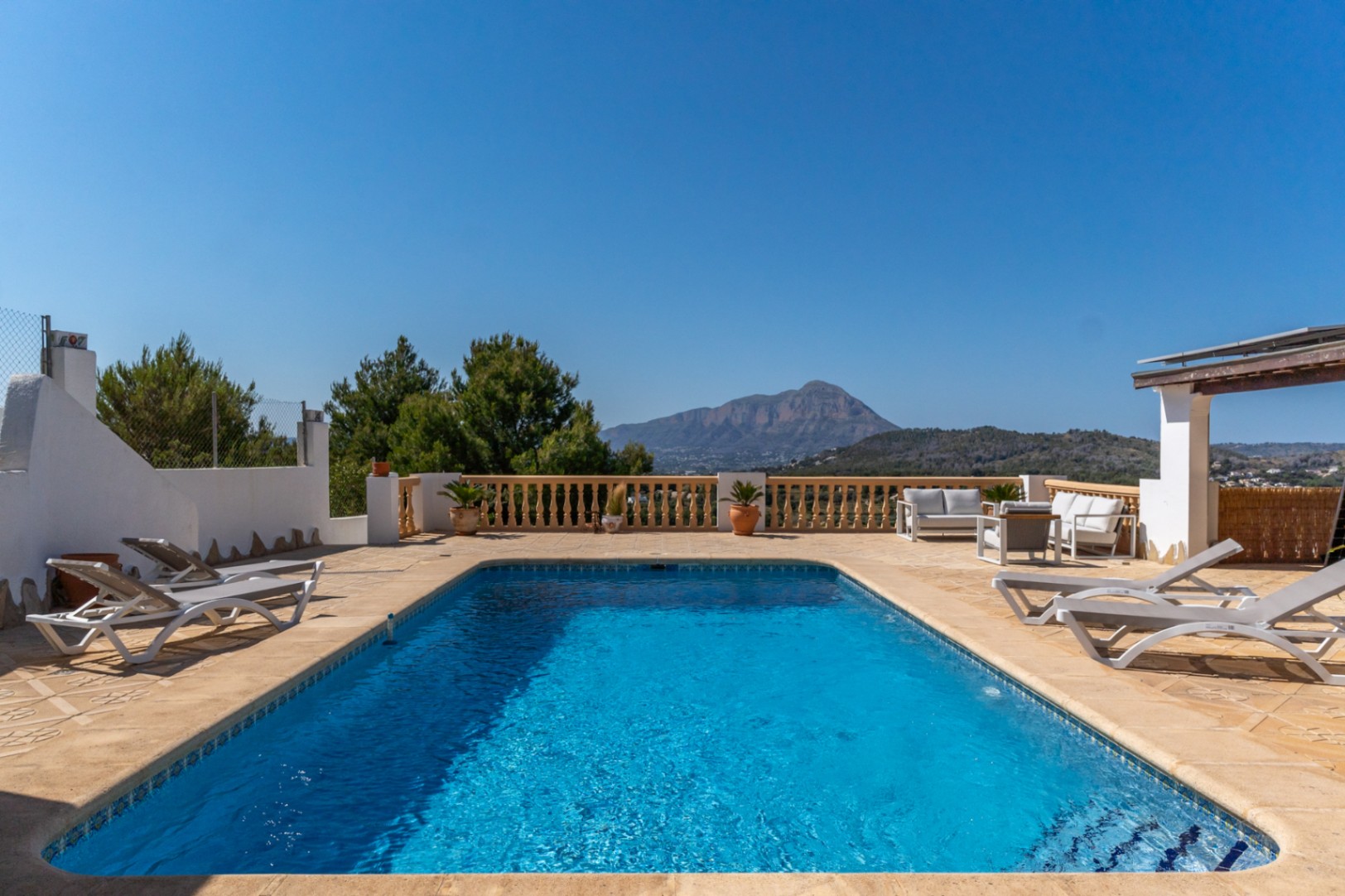 Rechteckiger Swimmingpool auf einer großzügigen Steinterrasse mit klassischer Balustrade und Blick auf den Montgó. Inklusive Sonnenliegen und einer komfortablen Lounge-Ecke.
