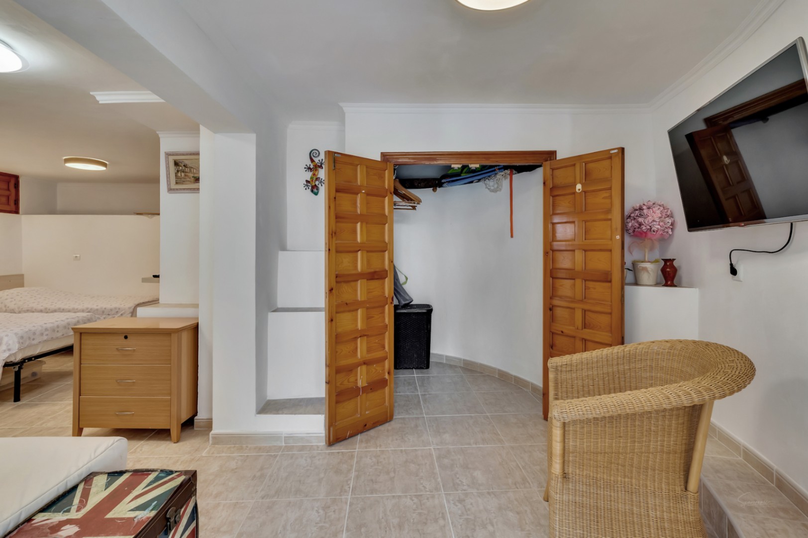 Schlafzimmer im Untergeschoss mit Fliesenboden, Einbauschrank aus Holz und Sitzbereich mit wandmontiertem Fernseher und Korbsessel.