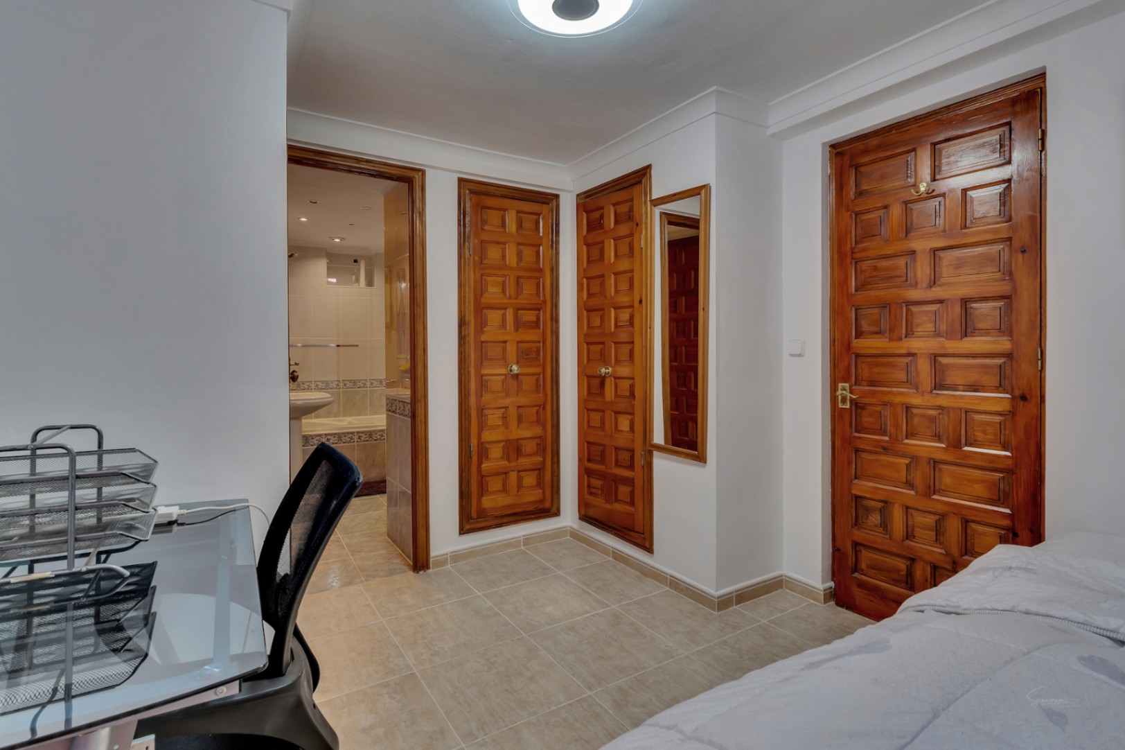 Schlafzimmer mit Einbauschränken aus Massivholz, Fliesenboden und direktem Zugang zum En-Suite-Badezimmer mit Badewanne und hellen Wandfliesen.