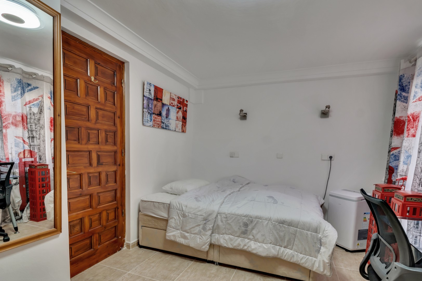 Schlafzimmer mit Fliesenboden, einer massiven Holztür mit Kassettenfüllung und Wandleuchten. Ein großer Spiegel und London-Dekoration prägen das helle und funktionale Ambiente.