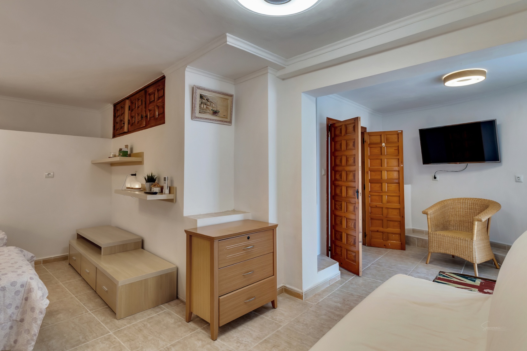 Schlafzimmer mit hellen Fliesenböden, Holzmöbeln, einem Korbsessel und traditionell geschnitzten spanischen Holztüren.