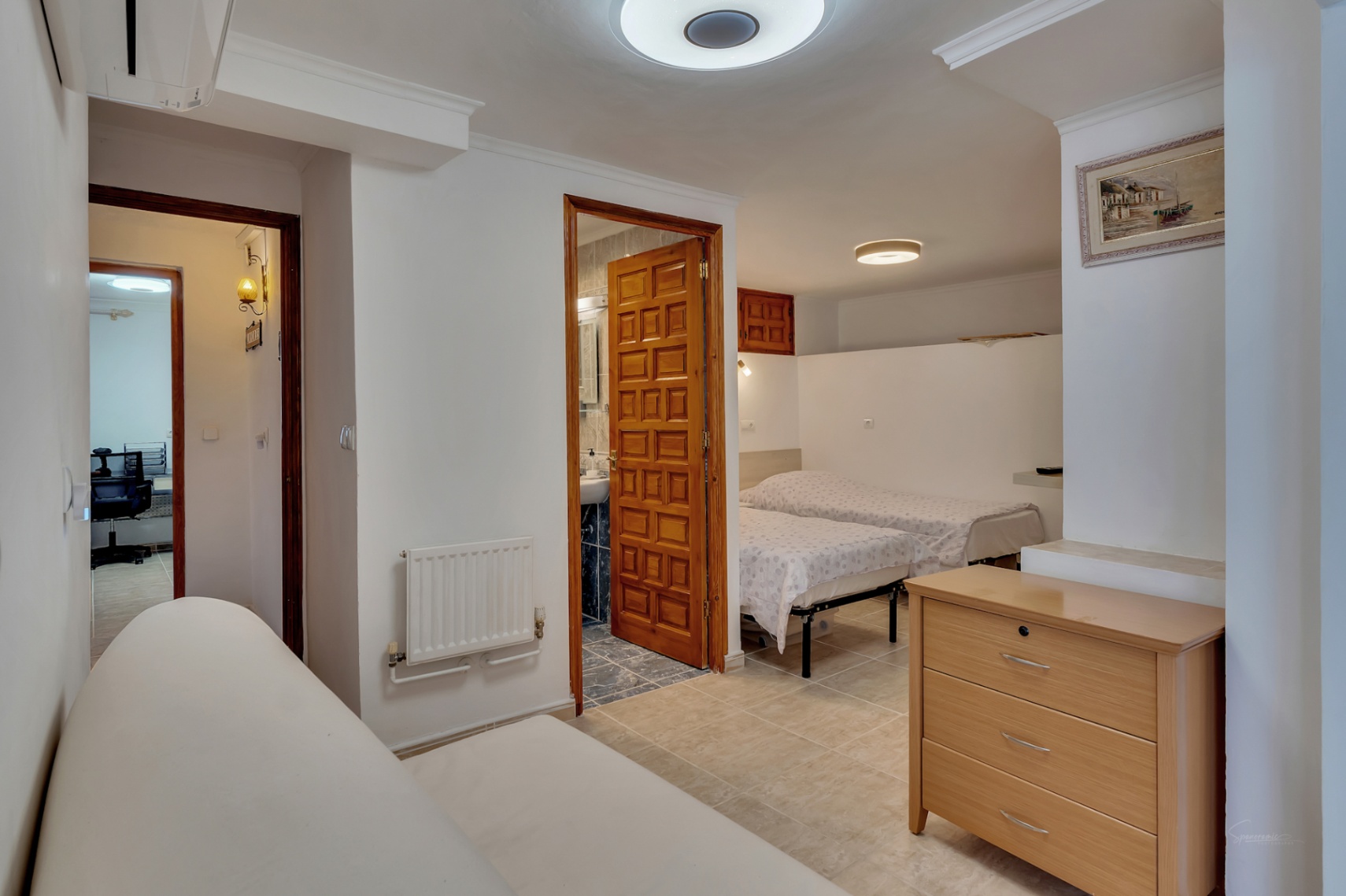 Schlafzimmer mit zwei Einzelbetten und En-Suite-Bad mit geschnitzter Holztür. Ausgestattet mit Wandheizkörper, Fliesenboden und Blick auf ein angrenzendes Büro.