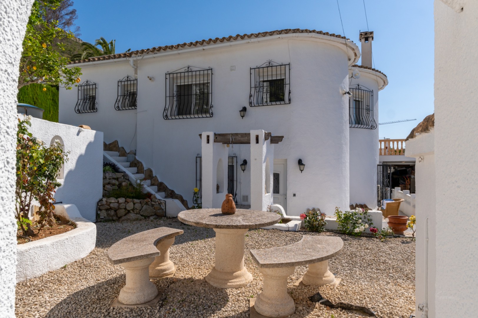 Weiße mediterrane Villa mit schmiedeeisernen Fenstergittern und einem Kieshof, der mit einem runden Steintisch und geschwungenen Bänken ausgestattet ist.