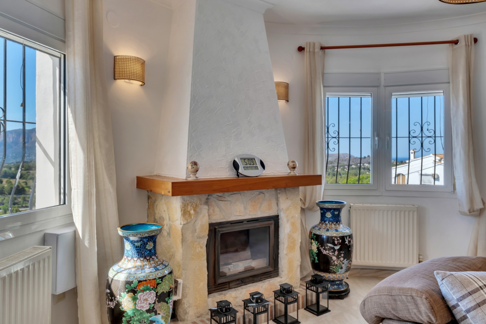 Wohnbereich mit Natursteinkamin und Holzsims, umgeben von Fenstern mit Blick auf den Montgó und das Meer sowie installierten Heizkörpern unter den Fenstern.