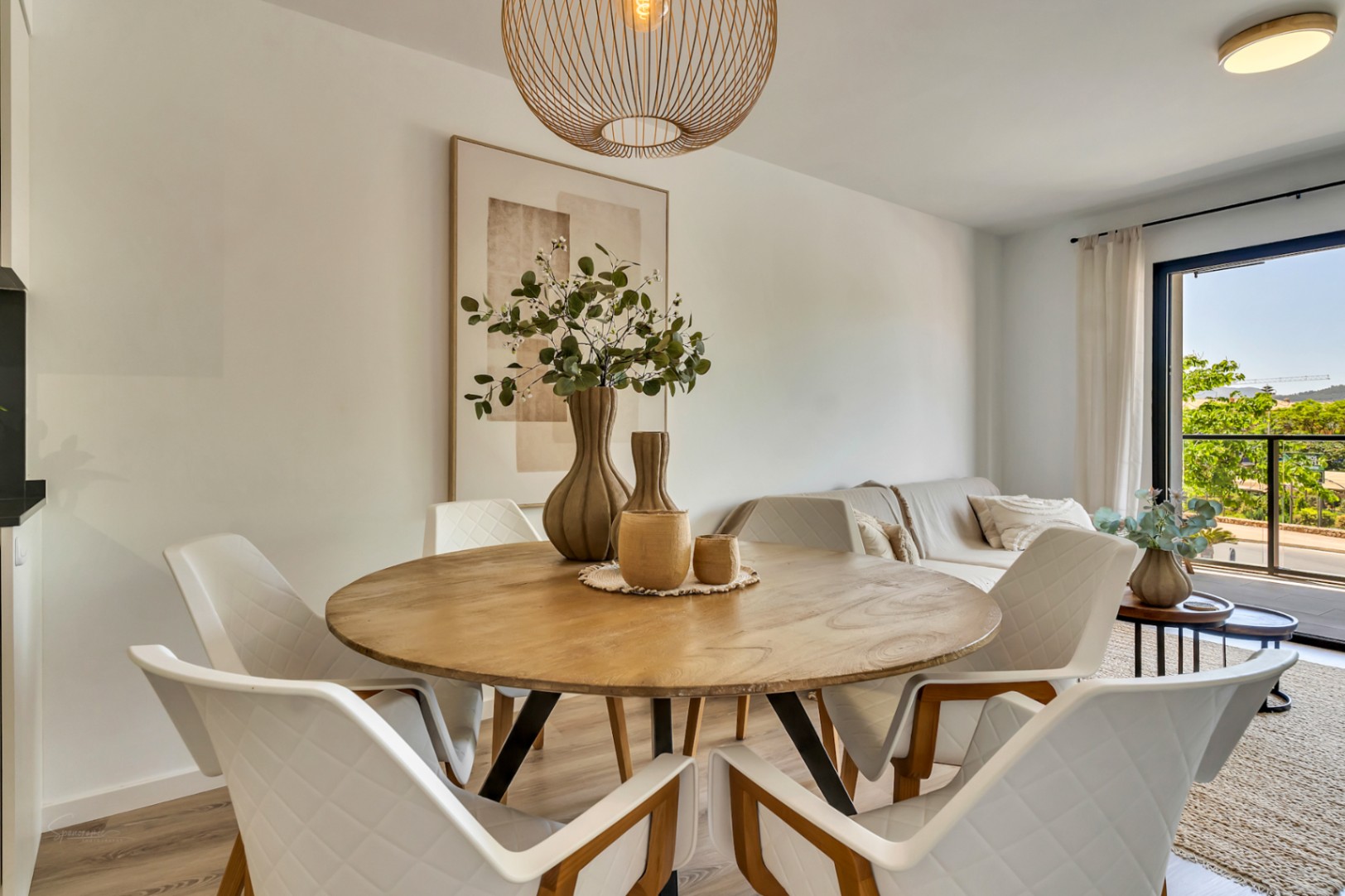 Essbereich mit rundem Holztisch und gesteppten Stühlen, beleuchtet durch eine geflochtene Pendelleuchte und Tageslicht von der angrenzenden Terrasse.