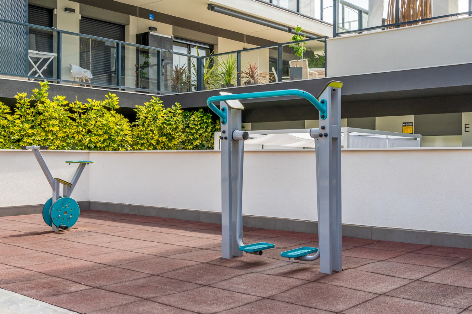 Fitnessbereich im Freien mit modernen Trainingsgeräten auf Fallschutzplatten, umgeben von Grünpflanzen und der zeitgenössischen Architektur der Anlage.