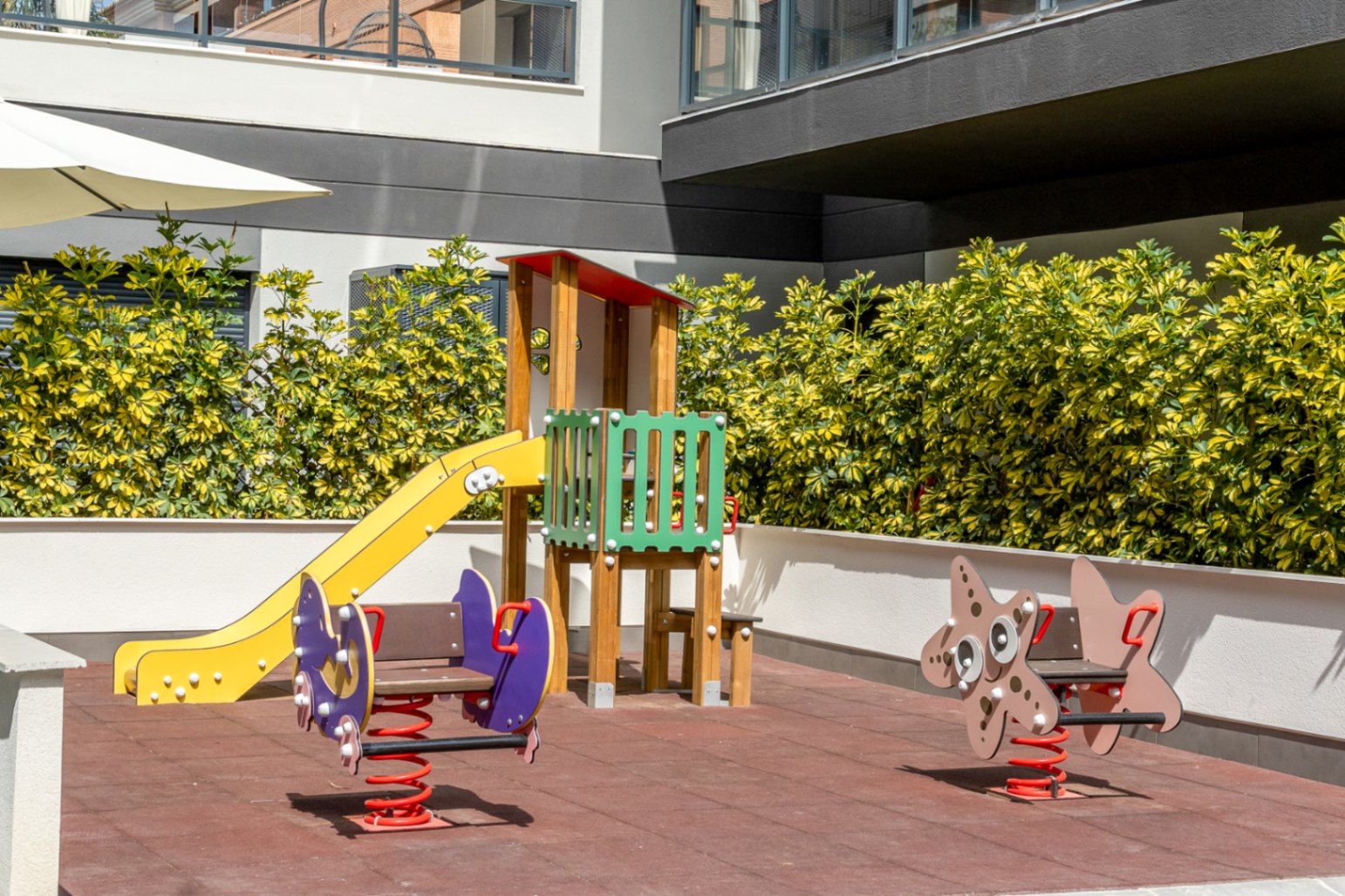 Gemeinschaftlicher Kinderspielplatz mit Fallschutzboden, gelber Rutsche und Federwippen, umgeben von gepflegten Hecken und moderner Wohnarchitektur.