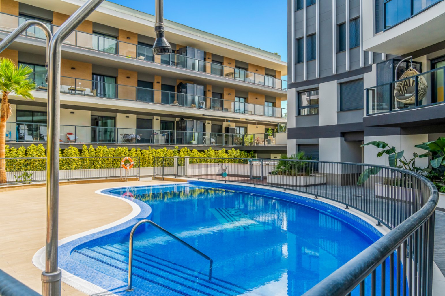 Gemeinschaftlicher Pool mit geschwungener Form, umgeben von einer Fliesenterrasse und modernen Wohngebäuden mit Glasbalkonen und Holzoptik.