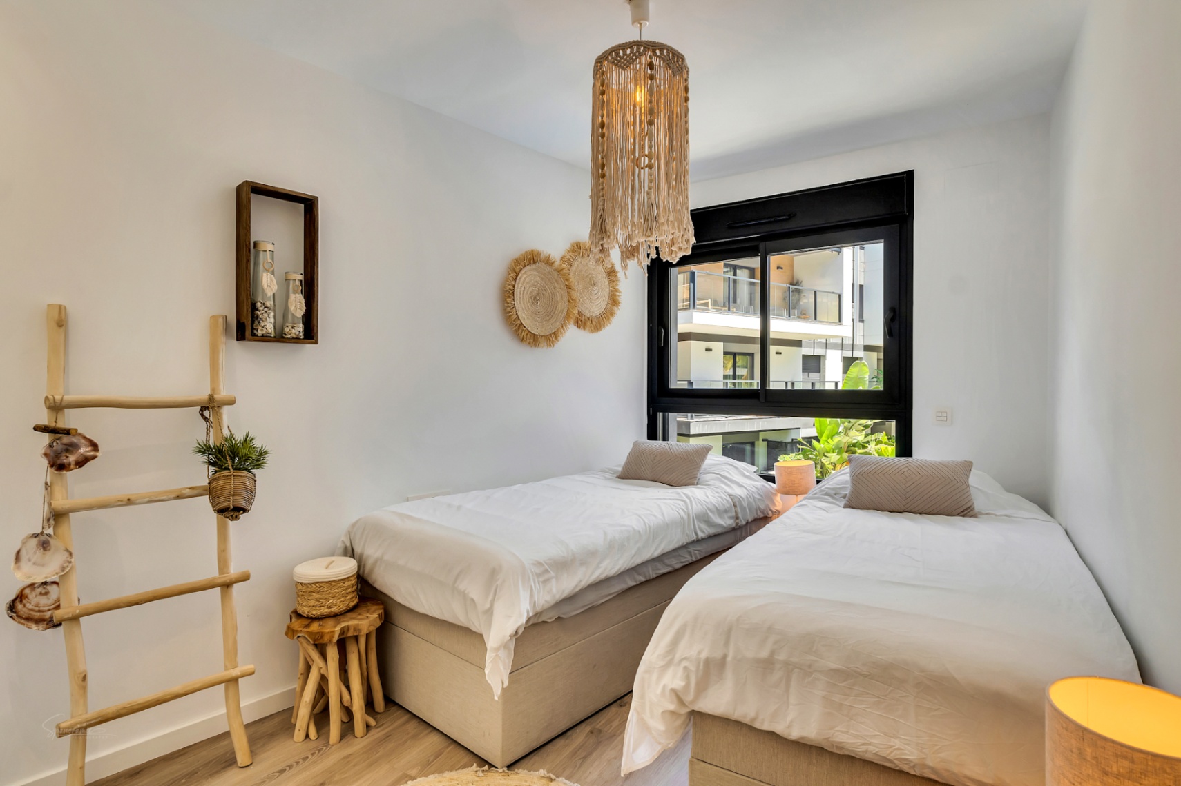 Helles Schlafzimmer mit zwei Einzelbetten, Akzenten aus Naturholz, einer Makramee-Pendelleuchte und einem schwarzen Fensterrahmen mit Blick auf die Anlage.