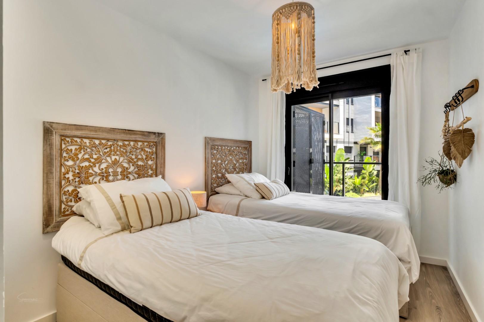 Helles Schlafzimmer mit zwei Einzelbetten, kunstvoll geschnitzten Holzkopfteilen und einer Makramee-Leuchte. Große Glasschiebetüren führen zur Terrasse mit Gartenblick.