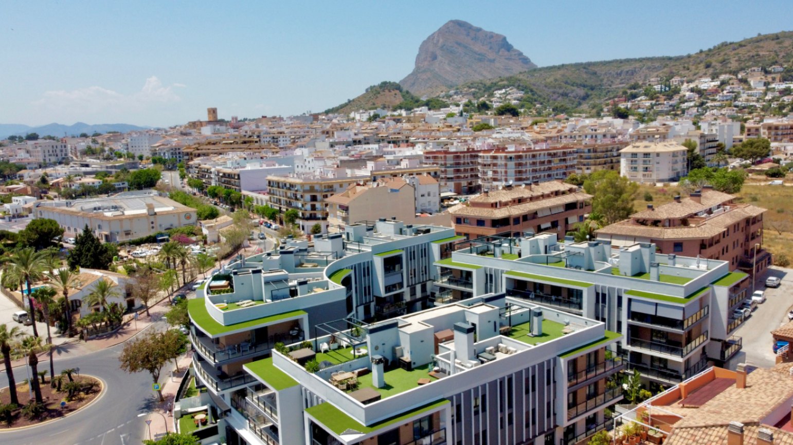Luftaufnahme der modernen Wohnanlage in Jávea mit Dachterrassen aus Kunstrasen und dem markanten Berg Montgó im Hintergrund unter blauem Himmel.
