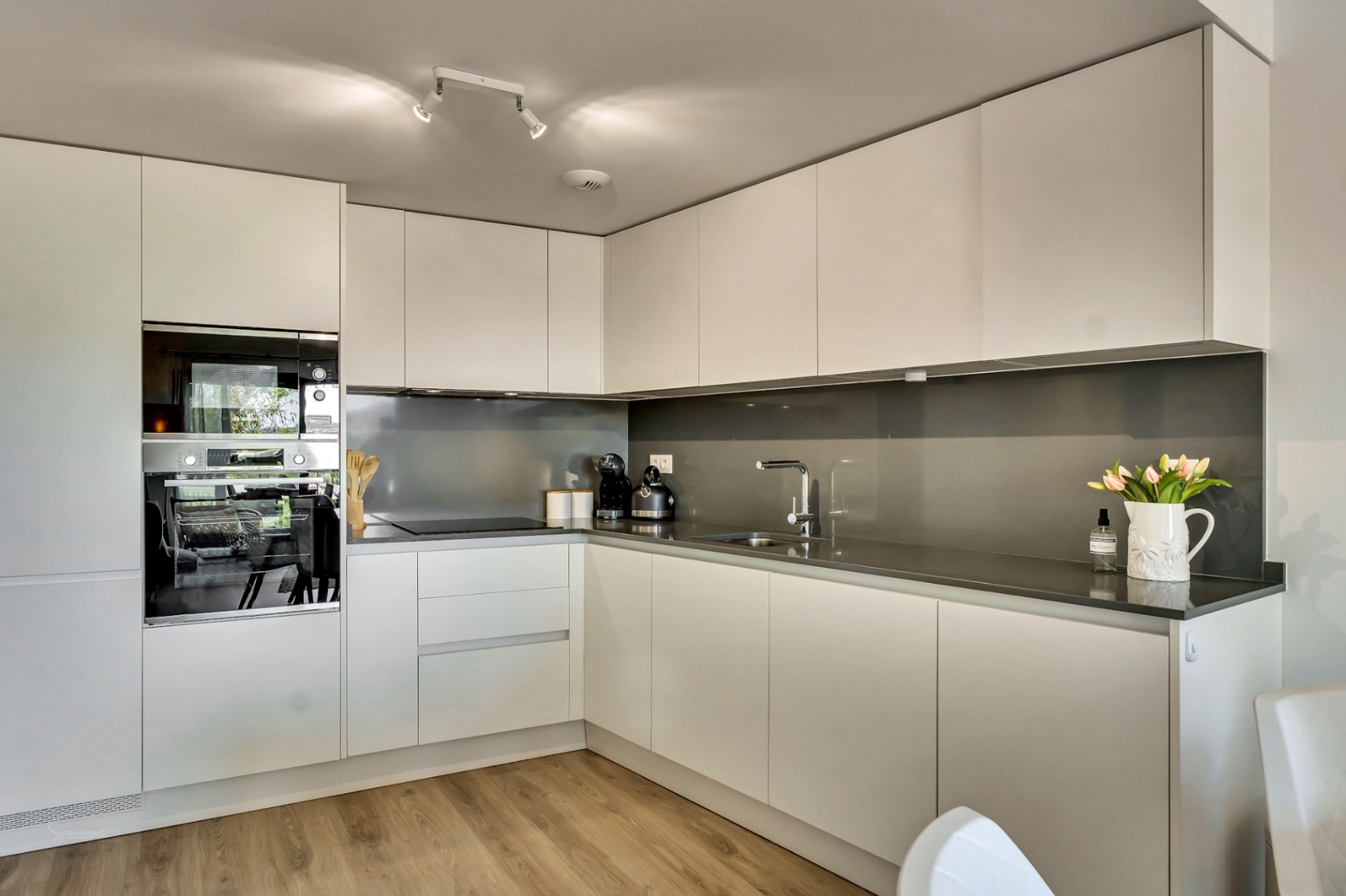 Moderne L-Küche mit minimalistischen weißen grifflosen Schränken, dunkelgrauer Arbeitsplatte und integrierten Einbaugeräten auf hellem Holzboden.