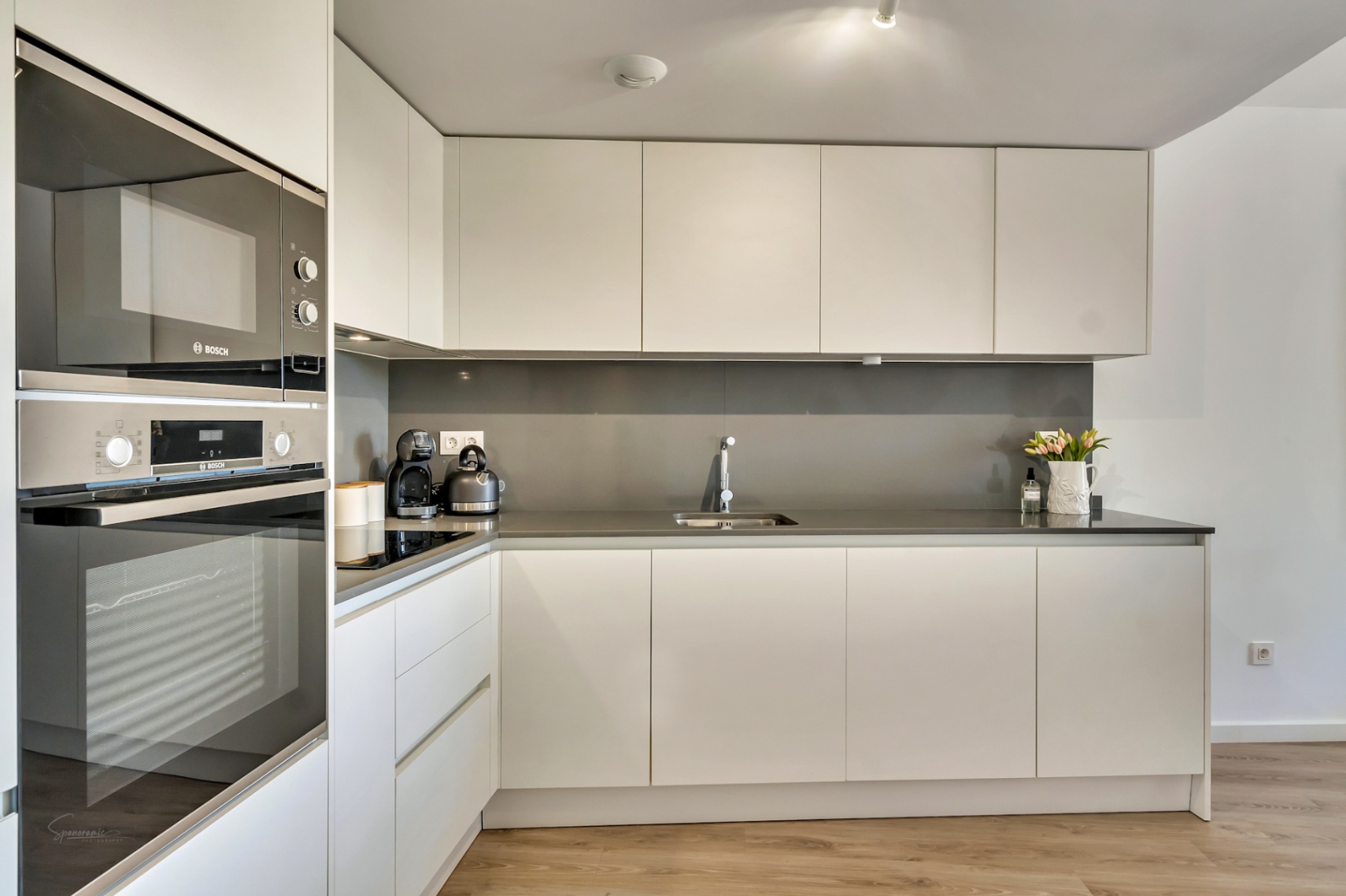 Moderne, minimalistische Küche mit grifflosen weißen Schränken, anthrazitfarbener Arbeitsplatte und integrierten Bosch-Einbaugeräten in einer Hochschrankzeile.