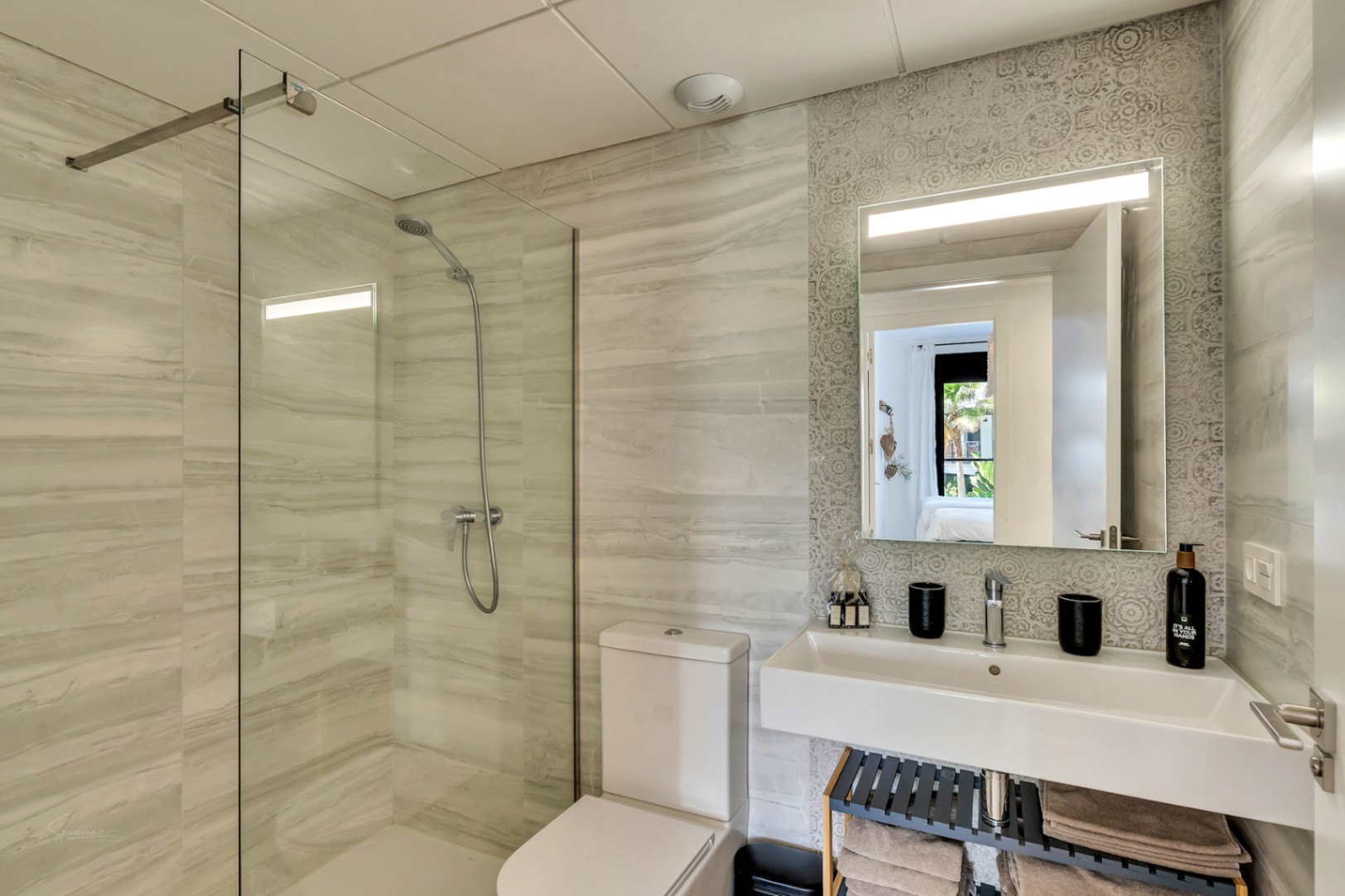 Modernes Badezimmer mit begehbarer Dusche, Glastrennwand und Fliesen in Marmoroptik. Ausgestattet mit einem Hängewaschbecken und einem beleuchteten Spiegel vor einer gemusterten Akzentwand.