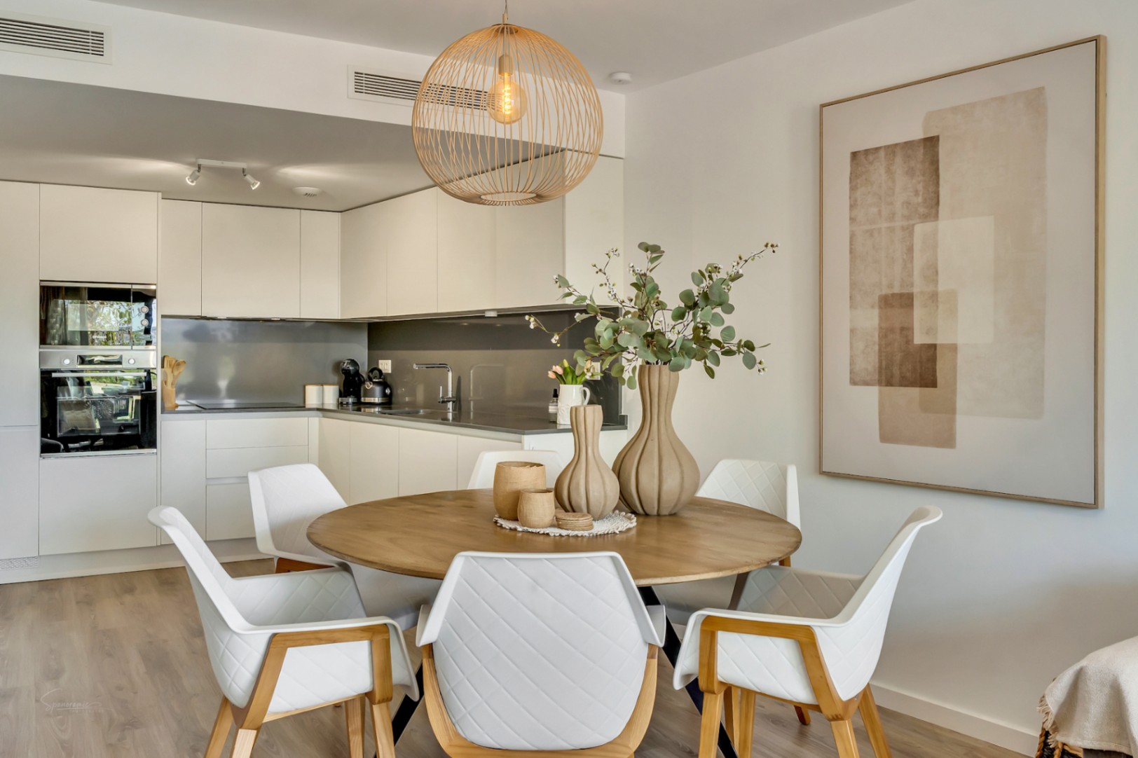 Offener Essbereich mit rundem Holztisch, gesteppten Design-Stühlen und einer Korblampe, direkt angrenzend an eine moderne, weiße Einbauküche.