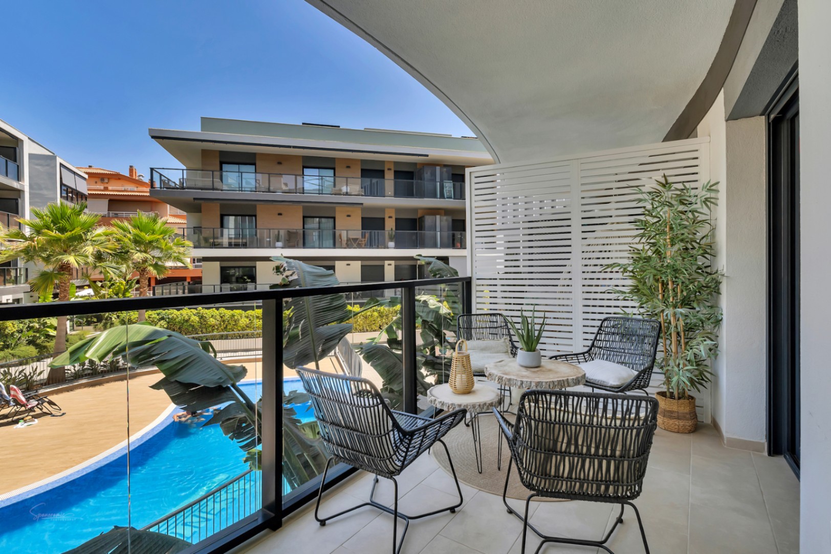 Private Terrasse mit Glasgeländer, modernen Gartenmöbeln und Blick auf den Gemeinschaftspool sowie die gepflegten Grünanlagen der Wohnanlage.