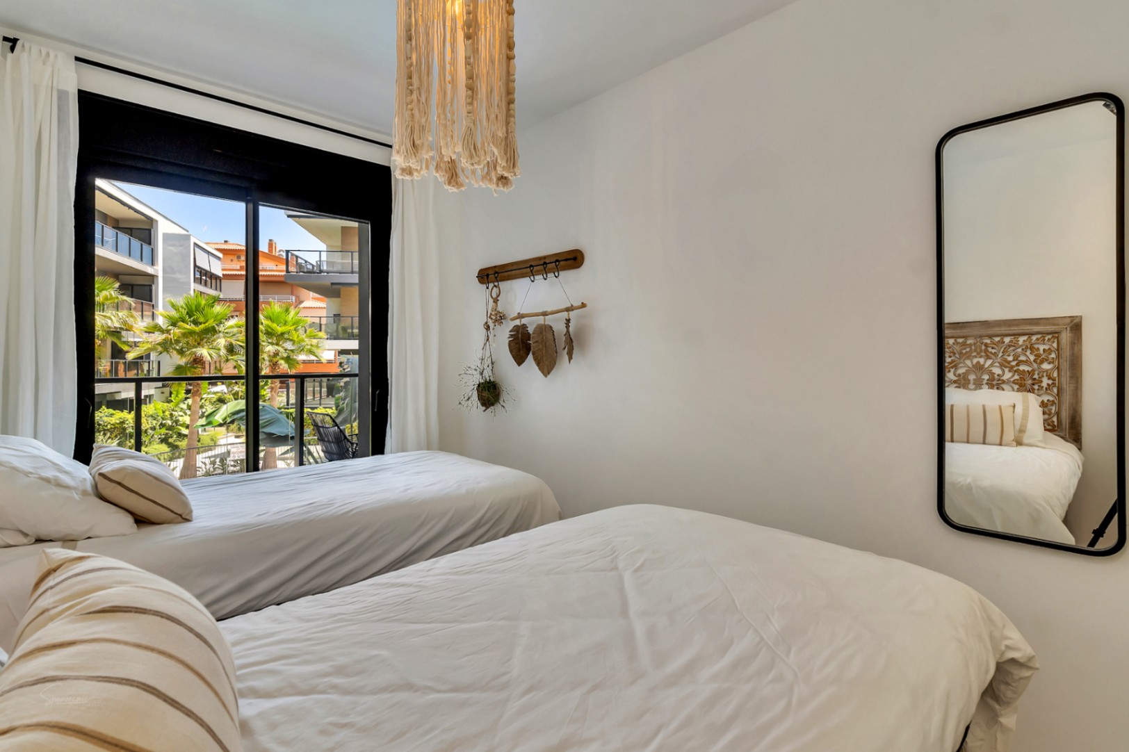 Schlafzimmer mit zwei Einzelbetten, Boho-Elementen und großen Glasschiebetüren, die einen Blick auf die gepflegte Gartenanlage und Palmen bieten.
