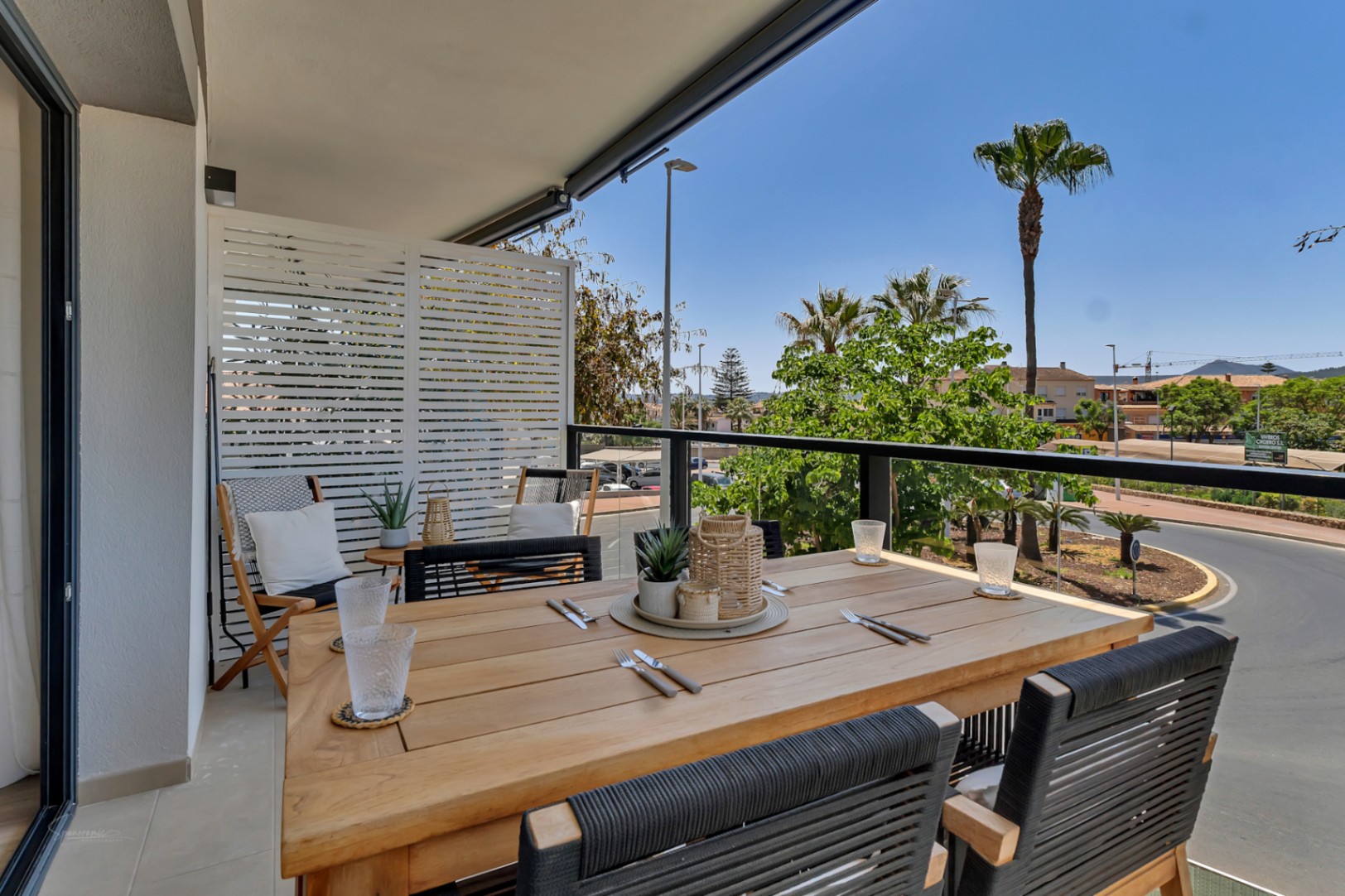 Überdachte Terrasse mit Holzesstisch, schwarzen Seilstühlen und freiem Blick auf die umliegenden Palmen und die Stadtlandschaft.