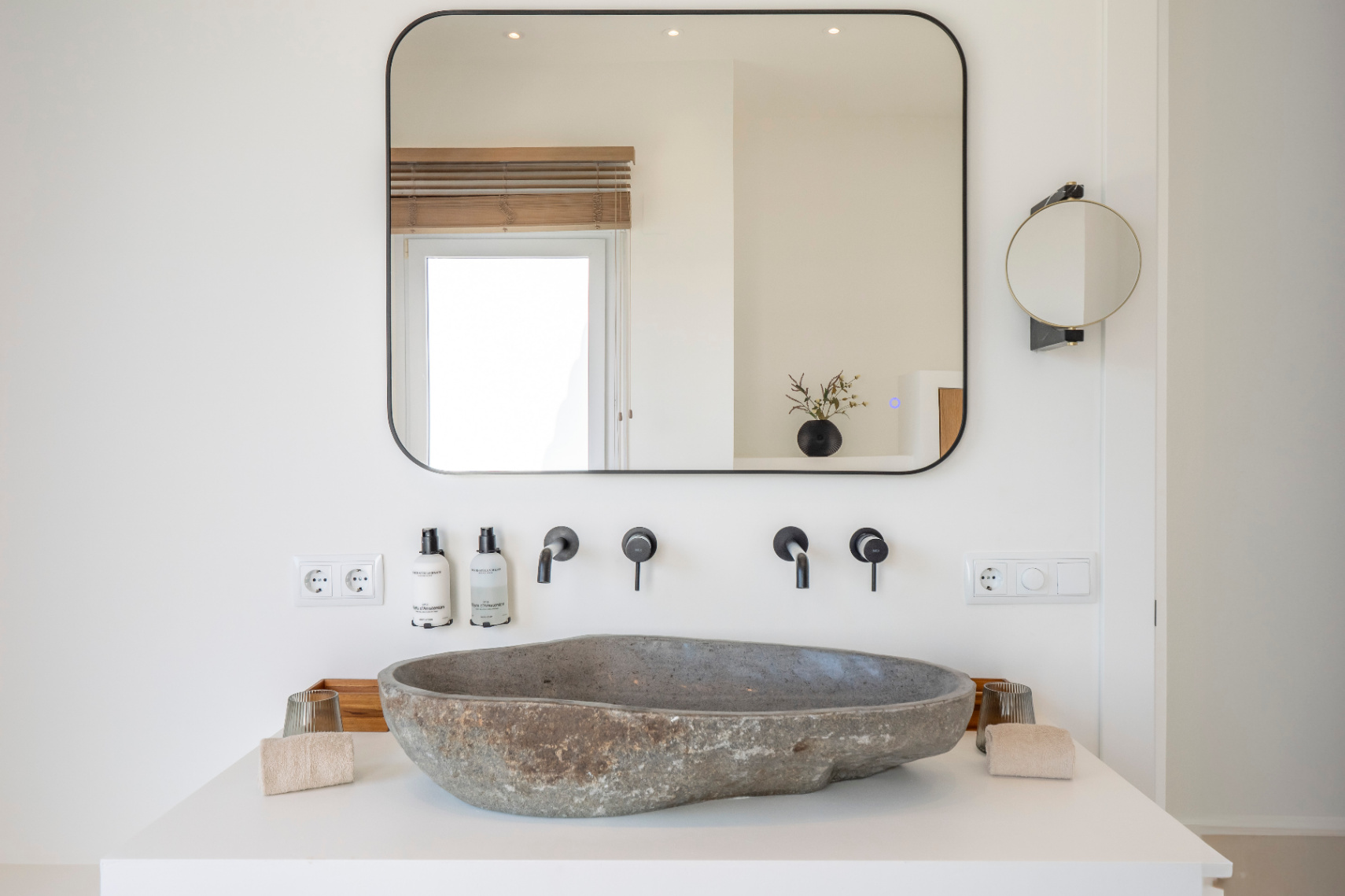 Badezimmerdetail mit Natursteinwaschbecken und mattschwarzen Armaturen. Rechteckiger Spiegel mit integrierter Beleuchtung.