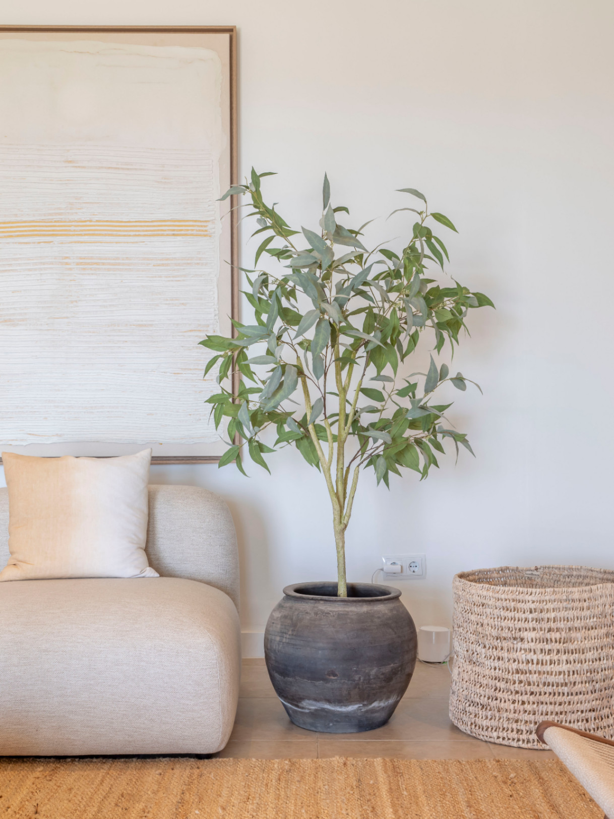 Detail des Wohnzimmers mit beigem Sofa, Topfpflanze aus Keramik und Weidenkorb auf einem Jute-Teppich.