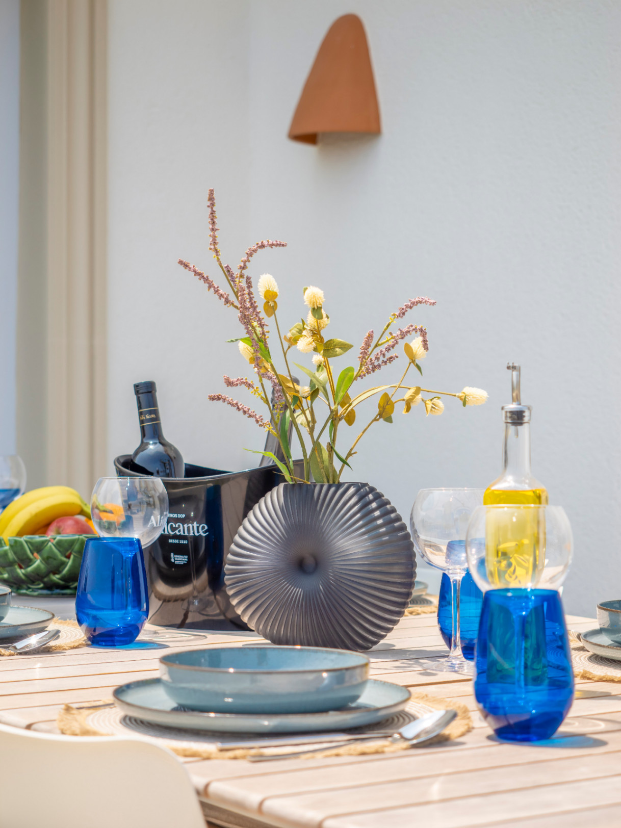 Esstisch im Freien mit blauem Geschirr, grauer Vase und Alicante-Weinkühler.