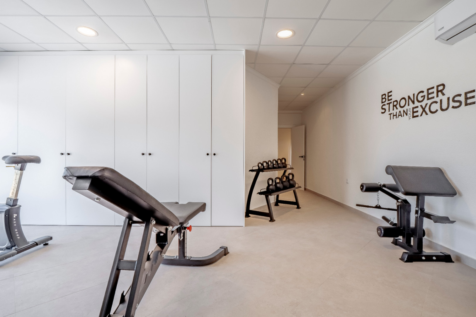 Fitnessraum mit Keramikböden, weißen Wänden und Einbauschränken. Trainingsgeräte mit Gewichten und Bank.