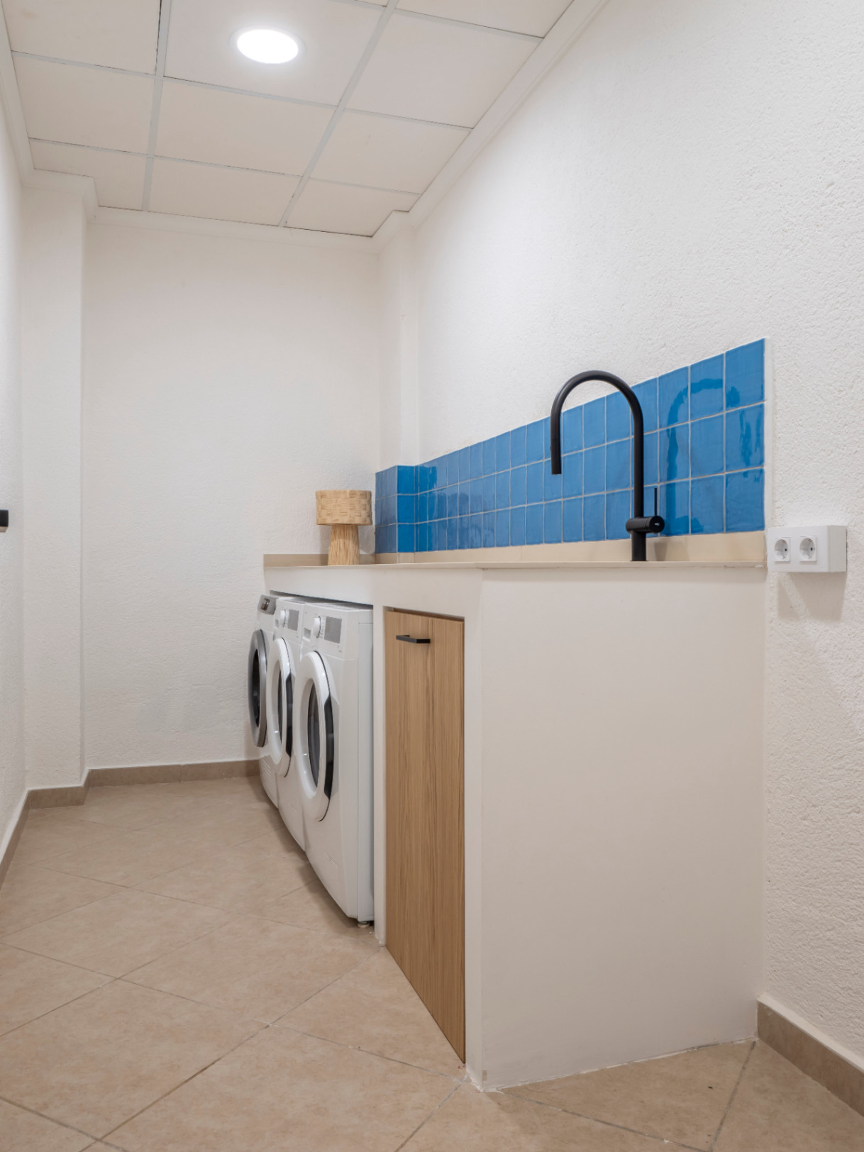 Funktionale Waschküche mit blauen Fliesen, beiger Arbeitsplatte, weißen Waschmaschinen und Fliesenboden.