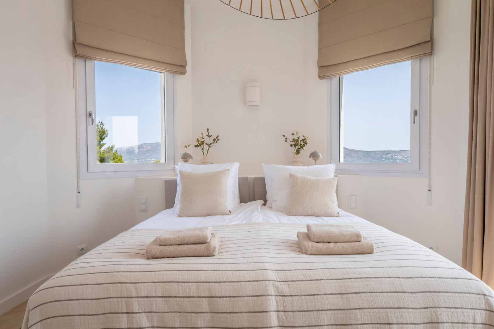 Helles Schlafzimmer mit zwei Fenstern mit Blick nach draußen. Doppelbett mit gestreifter Bettwäsche und gefalteten Handtüchern.