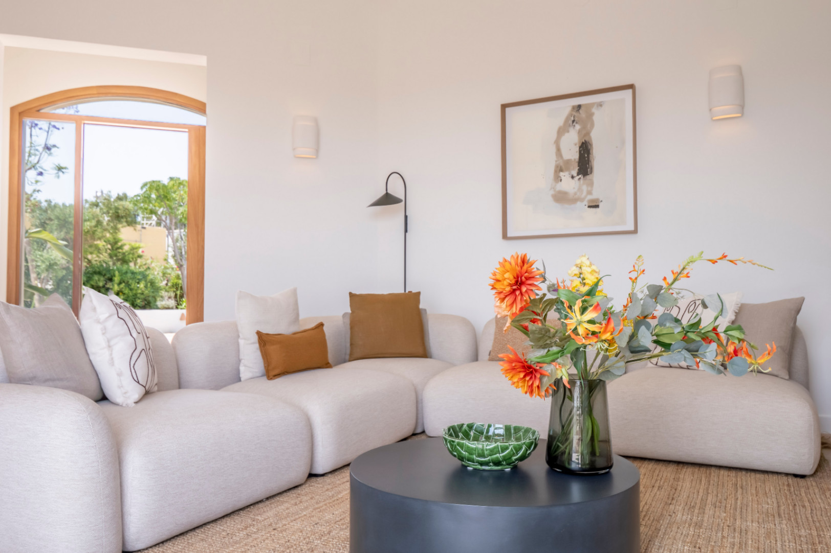 Helles Wohnzimmer mit beigem modularem Sofa, braunen Kissen und rundem schwarzem Tisch mit Blumendekoration.