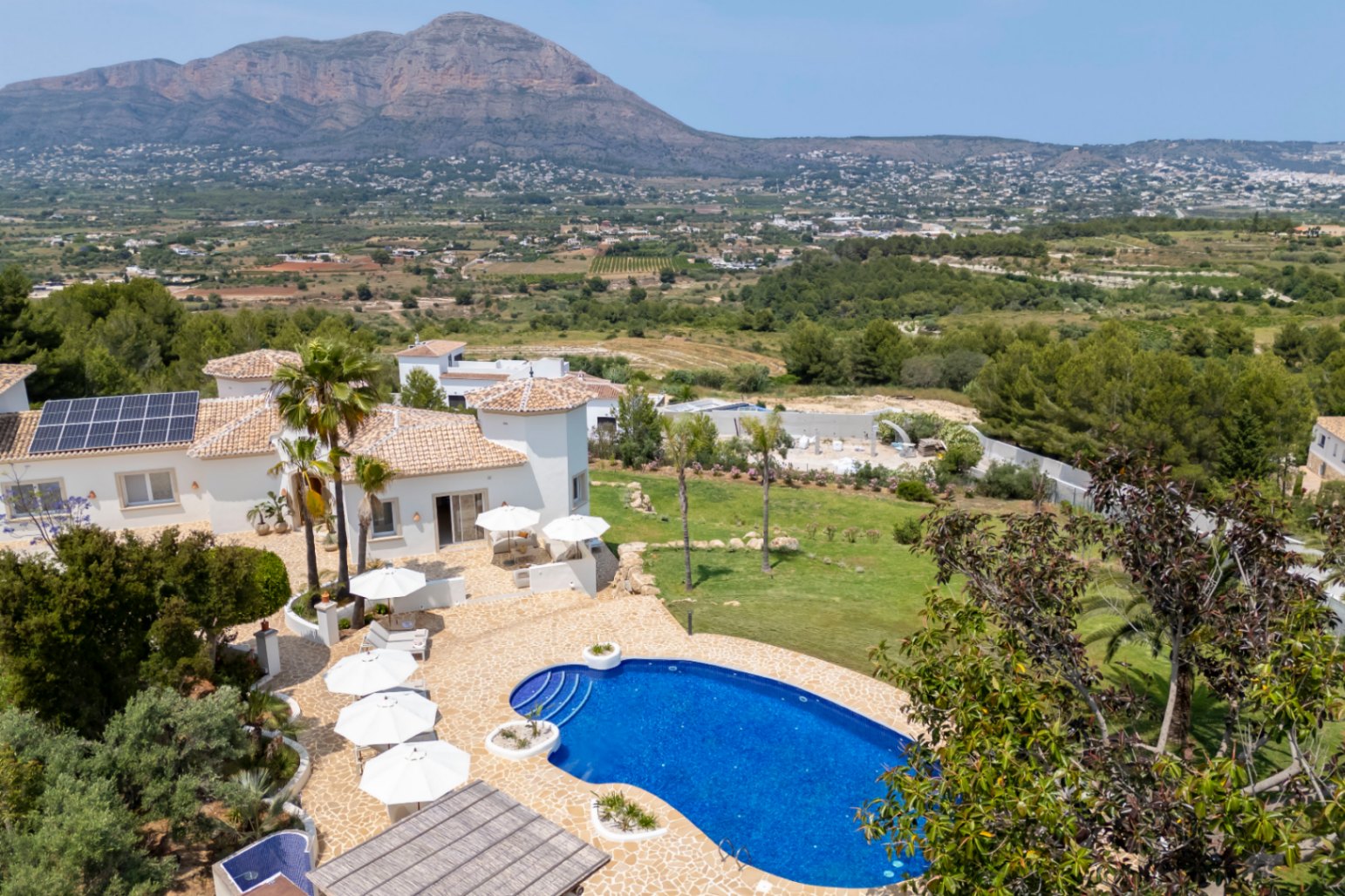 Luftaufnahme der Villa mit blauem Pool, Terrassen mit weißen Sonnenschirmen und mediterranem Garten mit Bergblick.