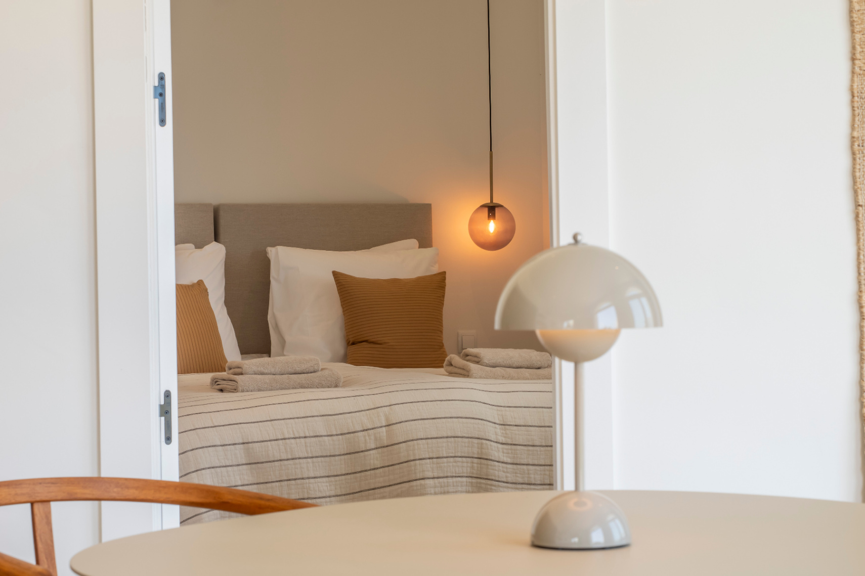 Schlafzimmer mit Doppelbett, kugelförmiger Pendelleuchte und gestreifter Bettwäsche. Runder Tisch mit Pilz-Tischlampe.