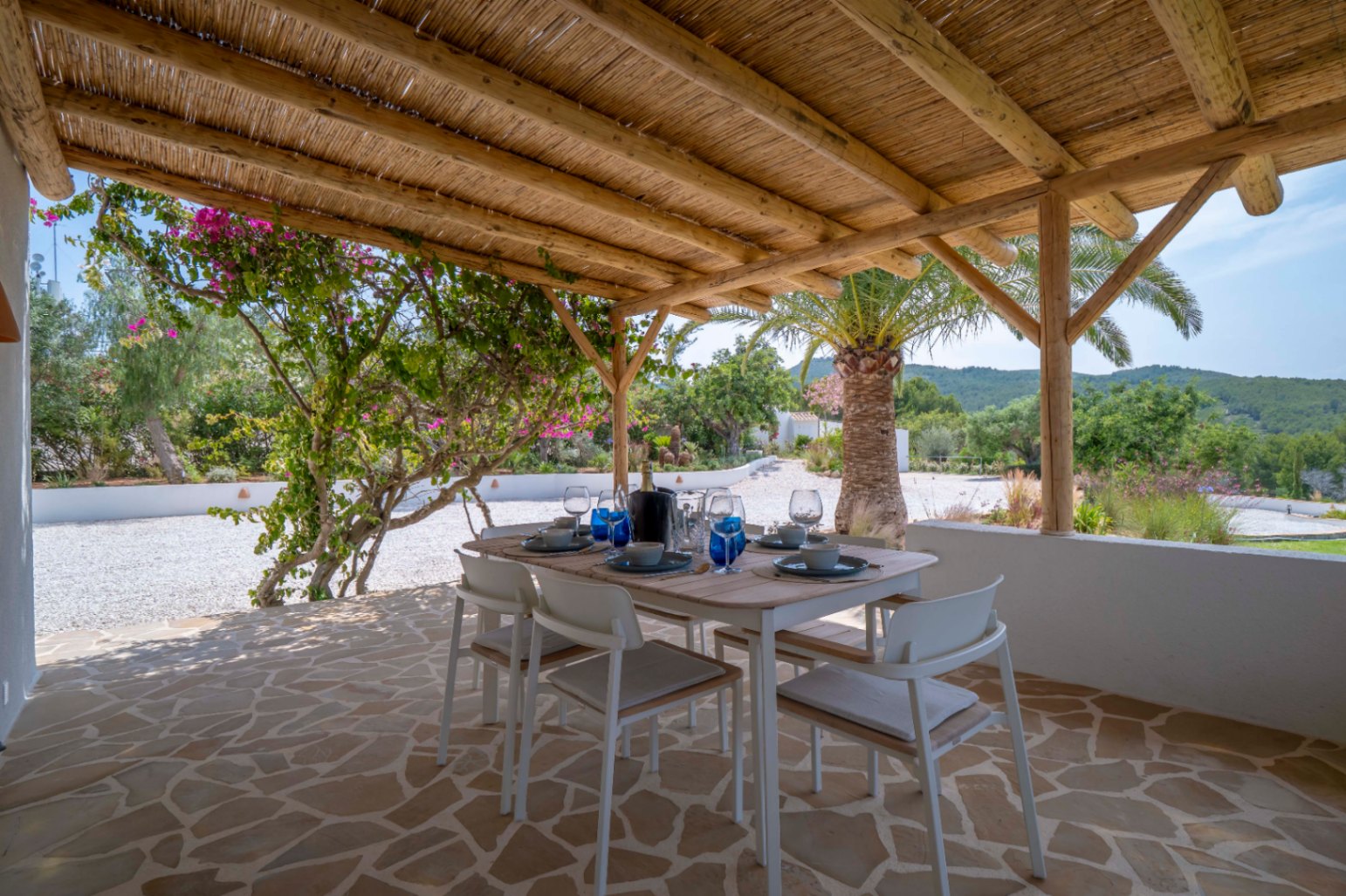 Überdachter Essbereich im Freien mit Strohdach, unregelmäßigem Steinboden und Blick auf den mediterranen Garten.