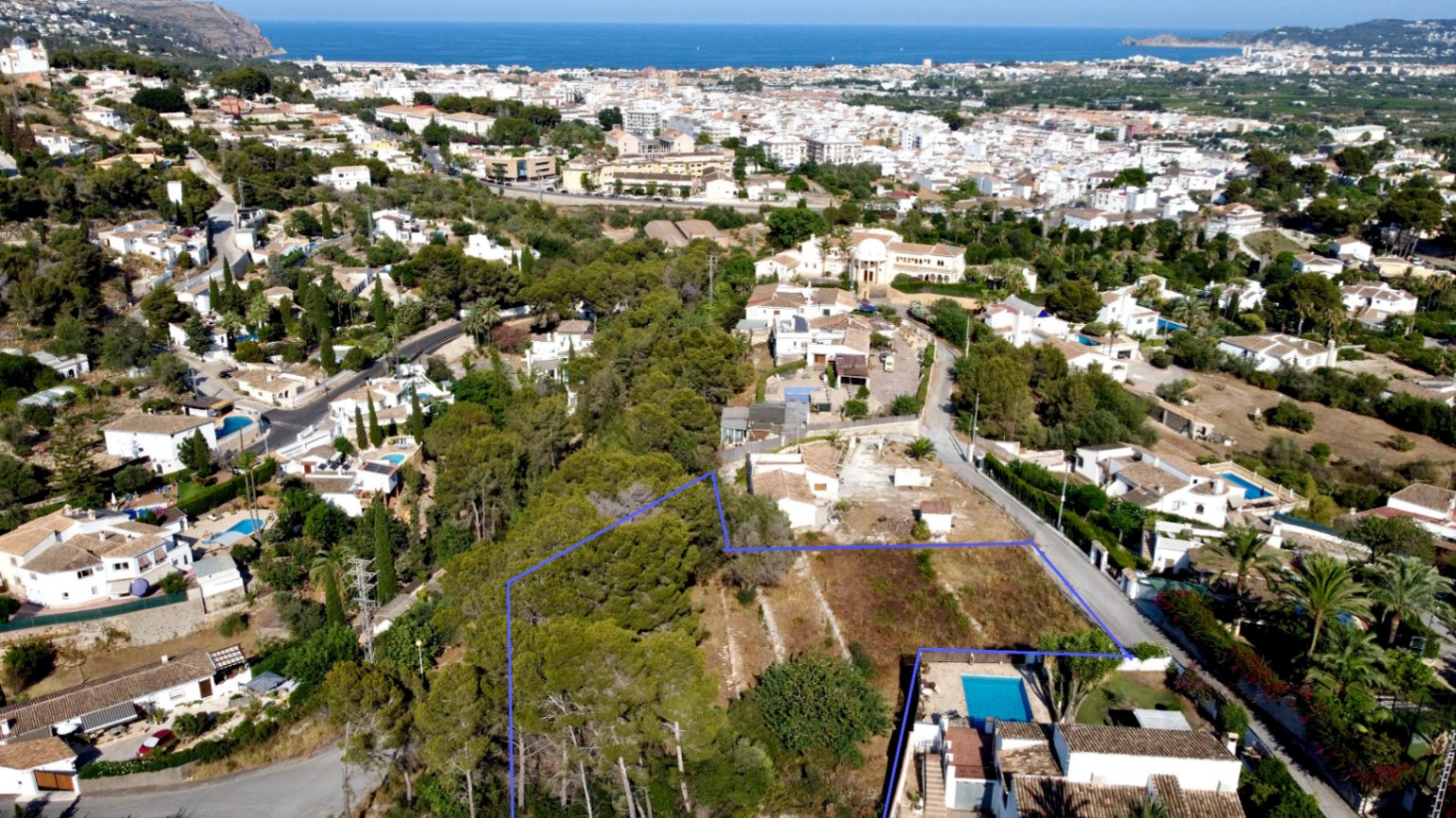 Luftaufnahme des 1.908 m² großen Grundstücks (blau markiert) mit Pinienbestand und Panoramablick auf das Meer und die Stadt Jávea.