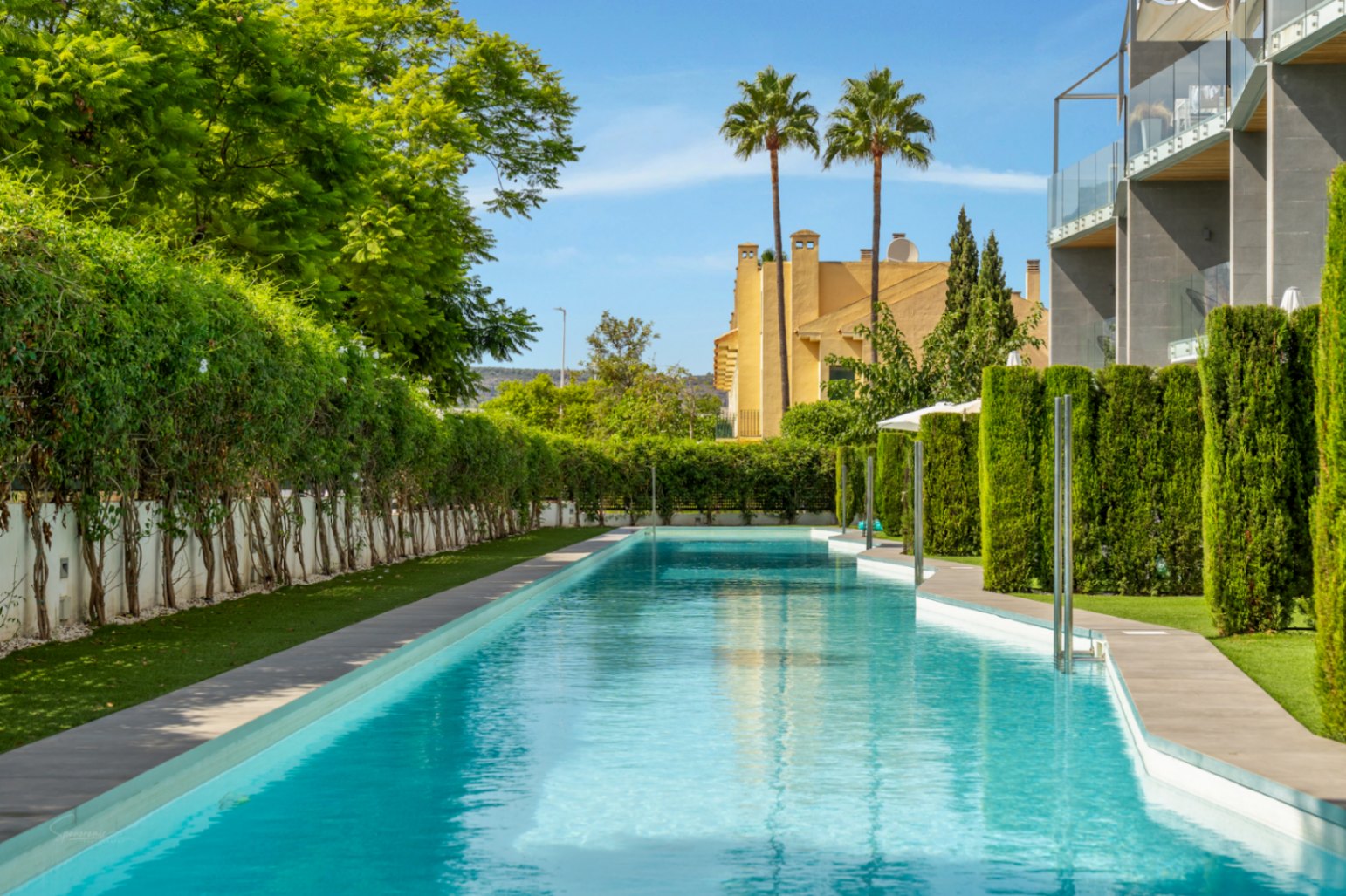 Ein 53 Meter langer Gemeinschaftspool mit türkisfarbenem Wasser, umgeben von gepflegten Hecken und moderner Architektur unter blauem Himmel.