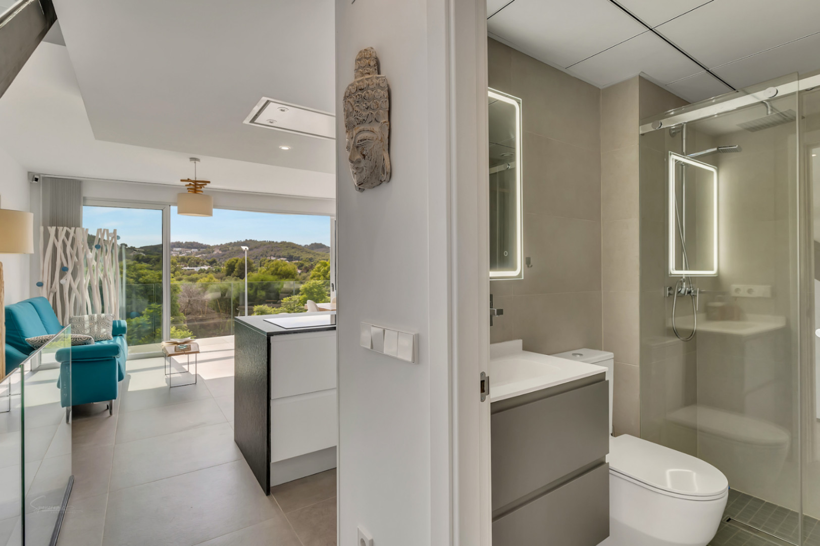 Flurblick auf ein modernes Badezimmer mit LED-Spiegel und Glasdusche sowie den offenen Wohnbereich mit weitem Blick auf die umliegende Landschaft.